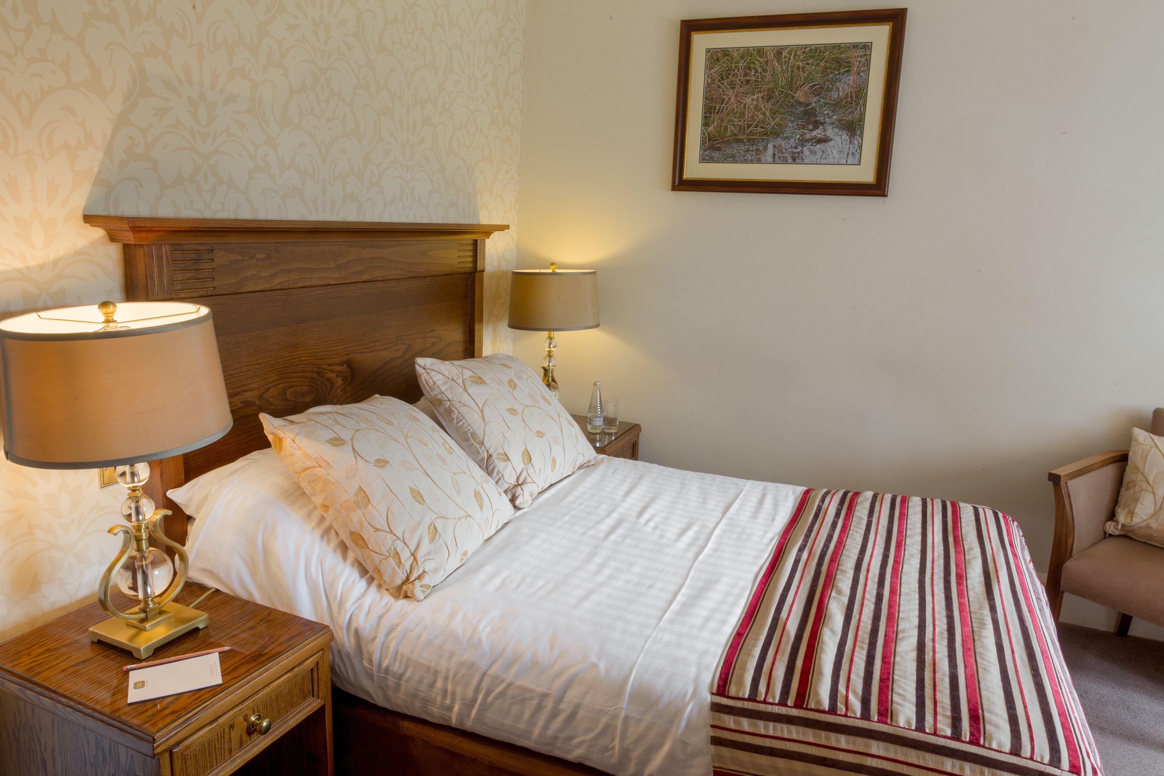 Una cama bien hecha con una manta a rayas, dos lámparas de noche, una mesita de noche de madera con una lámpara, un cuadro en la pared y una silla con cojín.