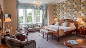 A bedroom with a large bed, a sitting area with a sofa and chairs, a desk with a lamp, and a window with curtains. - Classic Lodges Grinkle Park Easington (Saltburn-by-the-Sea)
