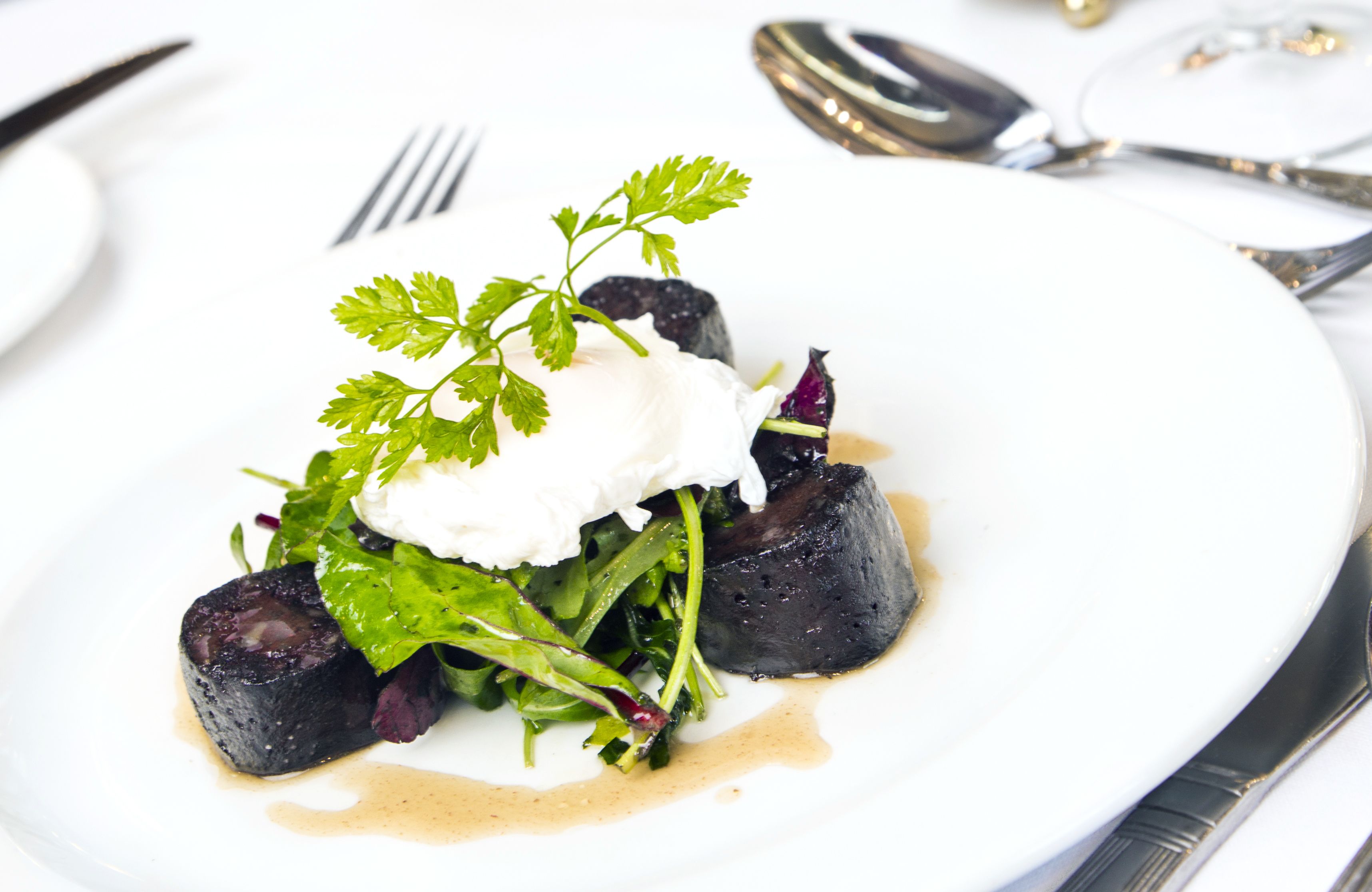 A white plate with a dark-colored food item, possibly a beetroot dish, garnished with greens and a dollop of cream.