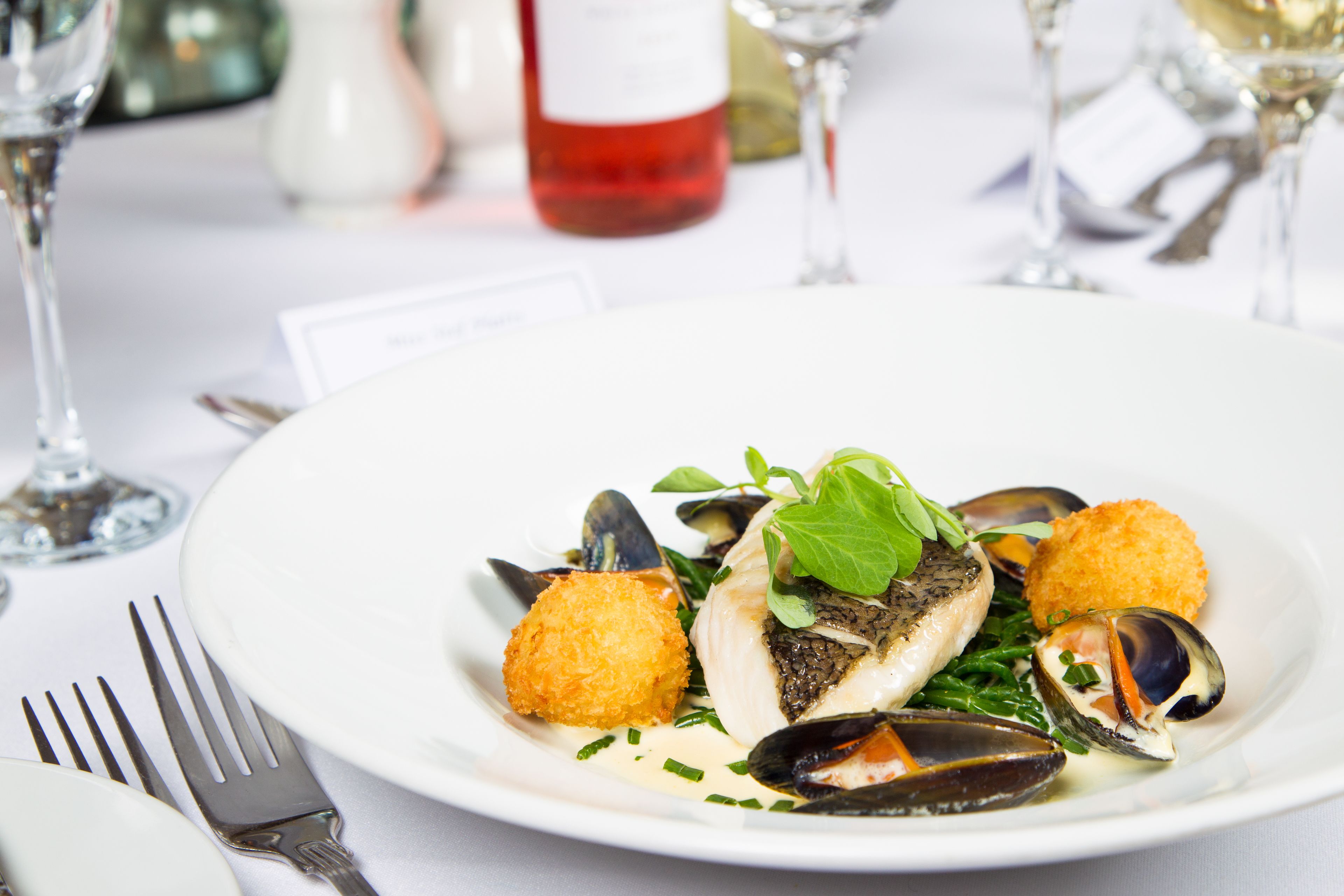 A plate of seafood with mussels, a fish fillet, and a garnish, accompanied by a fork and a wine glass.