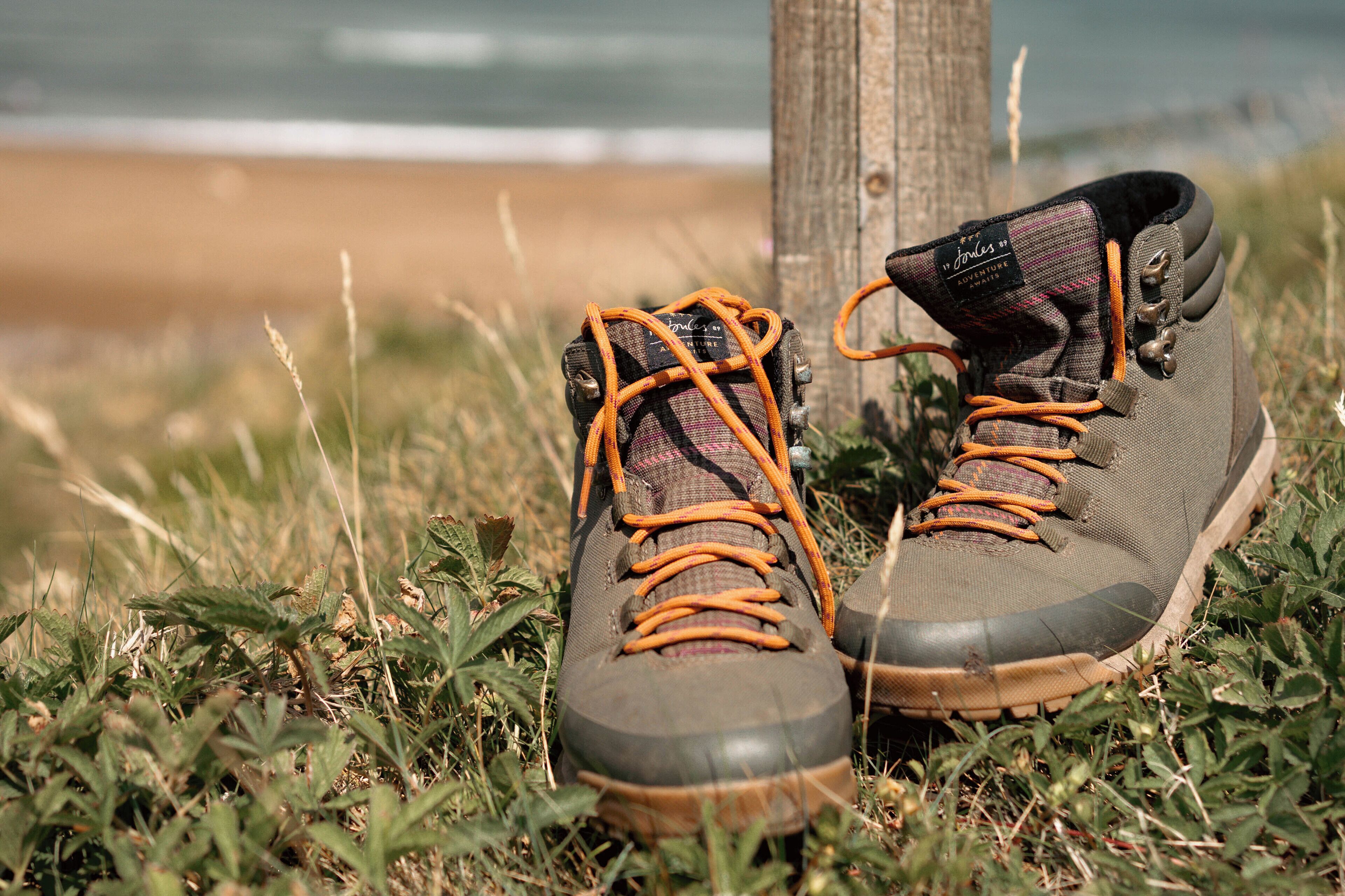 A pair of hiking boots with orange laces and a plaid sock, placed on grass near a wooden post.