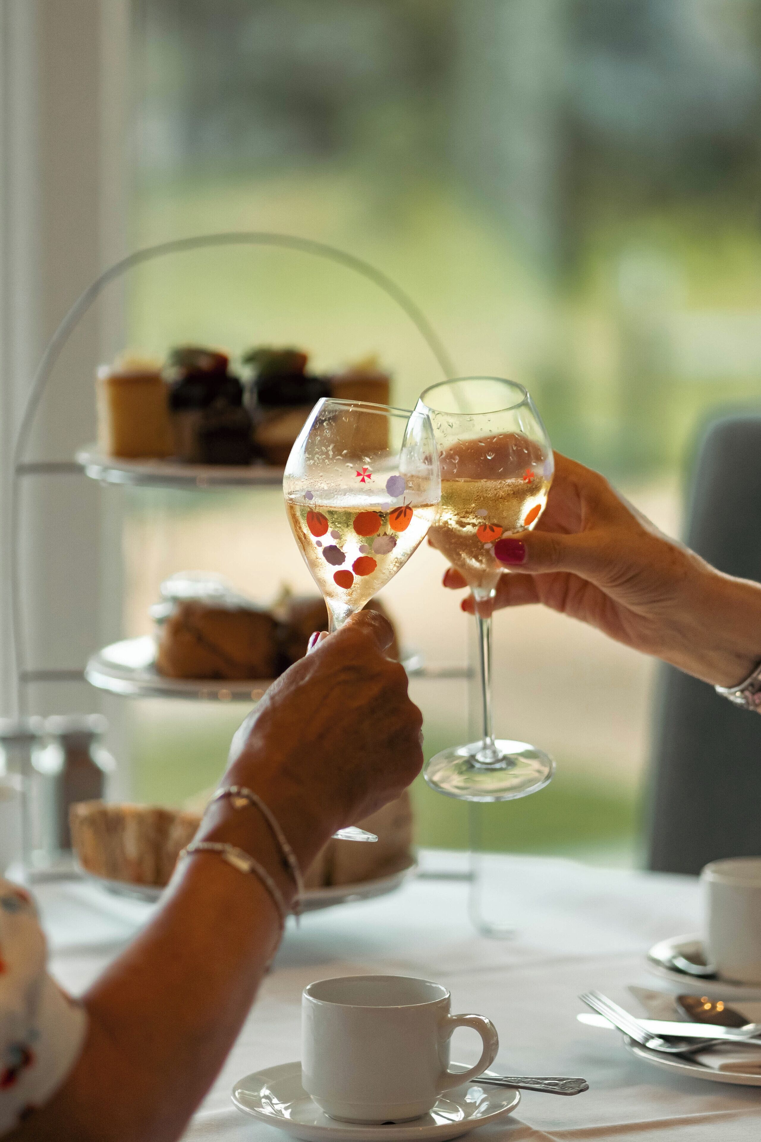 A table with a white cup and saucer, a glass of champagne, and a tiered dessert stand in the background.