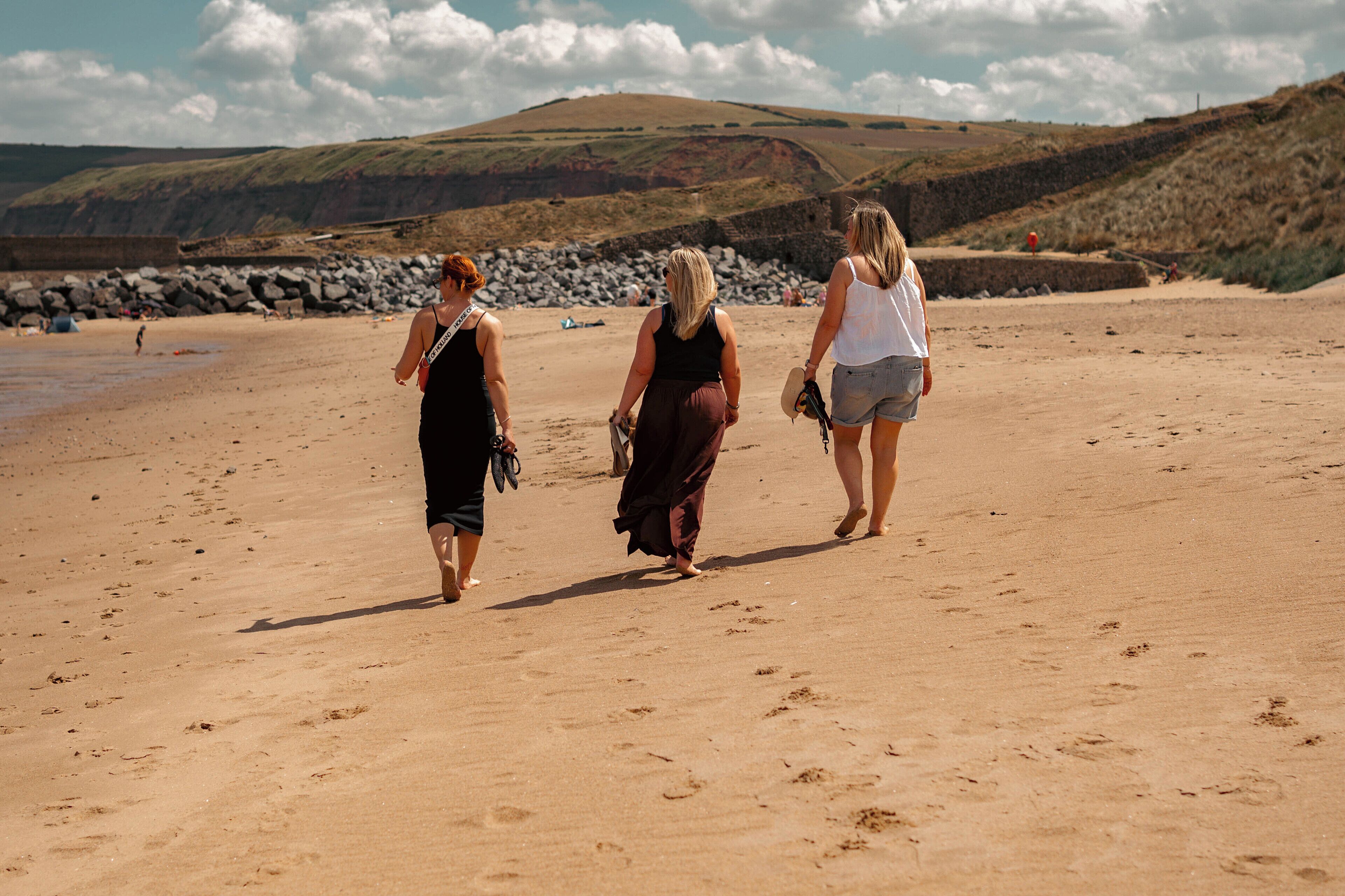 Drei Personen gehen über einen sandigen Strand, im Hintergrund steilen Felsen.