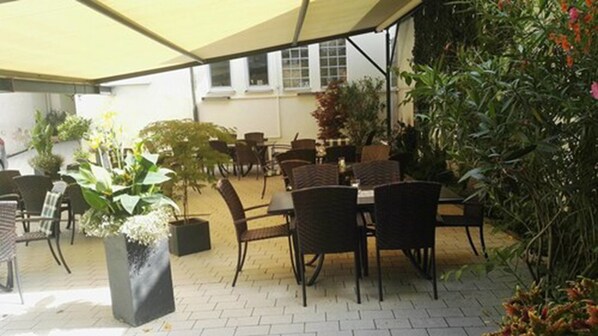 An outdoor patio area with tables and chairs, surrounded by potted plants and a large umbrella.