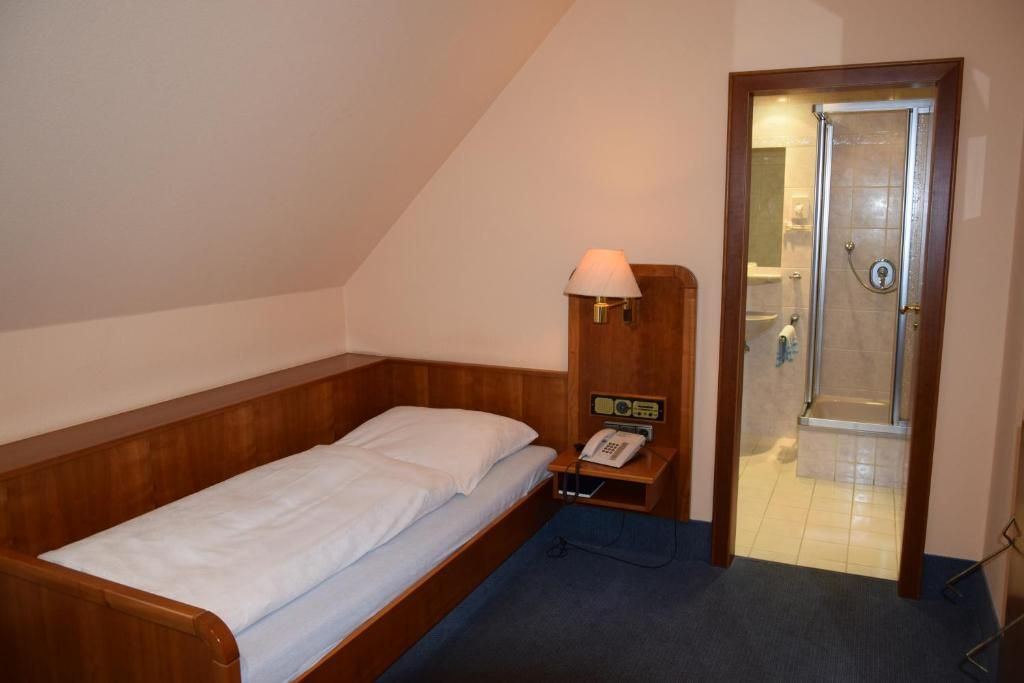 A single bed with a wooden headboard, a bedside telephone, a lamp, and a door leading to a bathroom with a shower.