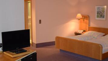 A hotel room with a bed, a TV on a wooden stand, a bedside table with a lamp, and a painting on the wall.