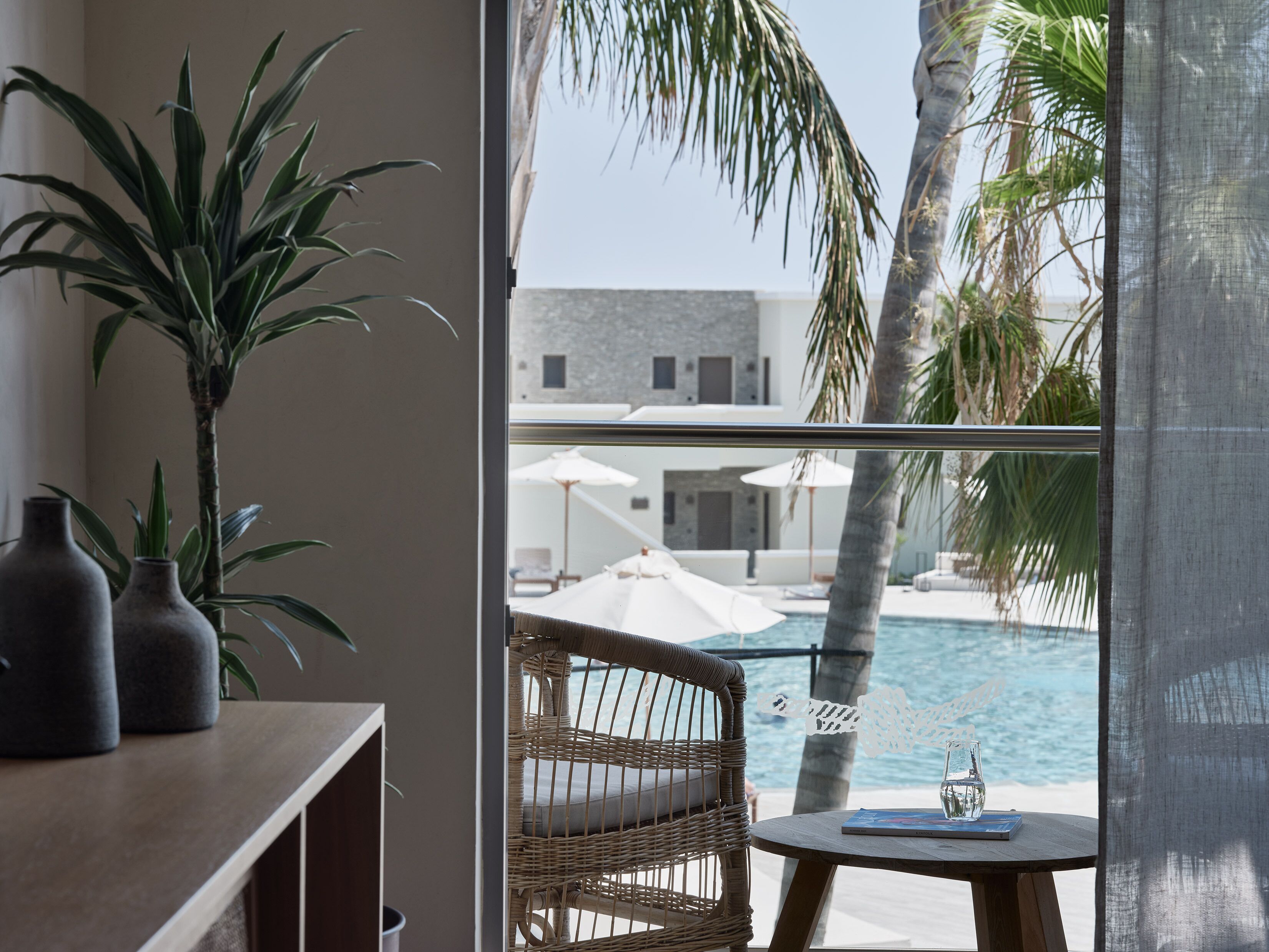 Un balcón con una silla de mimbre, una mesita y vista a una piscina y palmeras.