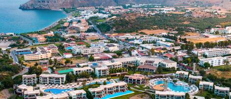 An aerial view of a coastal resort with multiple hotel buildings, a large swimming pool, and a beach area.