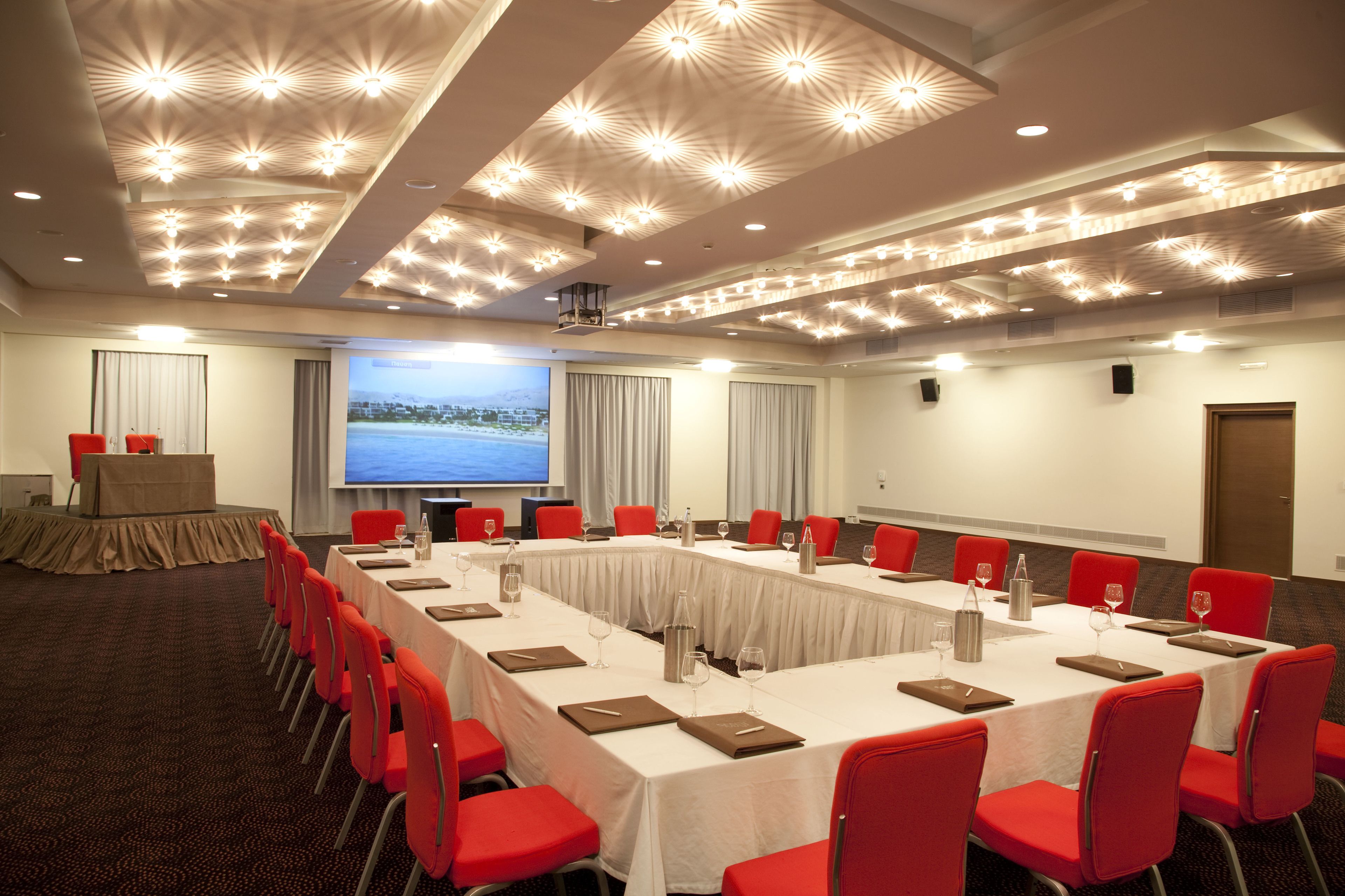 Un salón de conferencias con una larga mesa, sillas rojas y una pantalla grande.