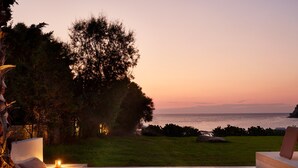 A modern outdoor seating area with a fire pit, overlooking a grassy landscape and the sea at sunset. - Port Royal Villas & Spa (Rhodes)