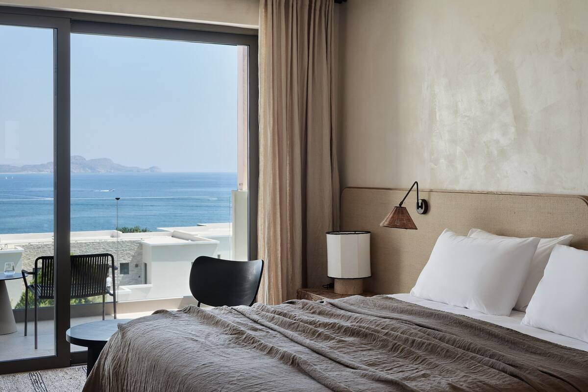 Une chambre d’hôtel avec un lit, une chaise, une table, une lampe et une vue sur la mer.