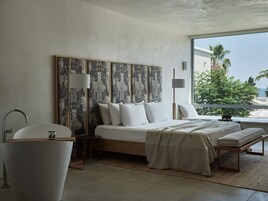 A modern bedroom with a large bed, a bathtub, and a view of palm trees.