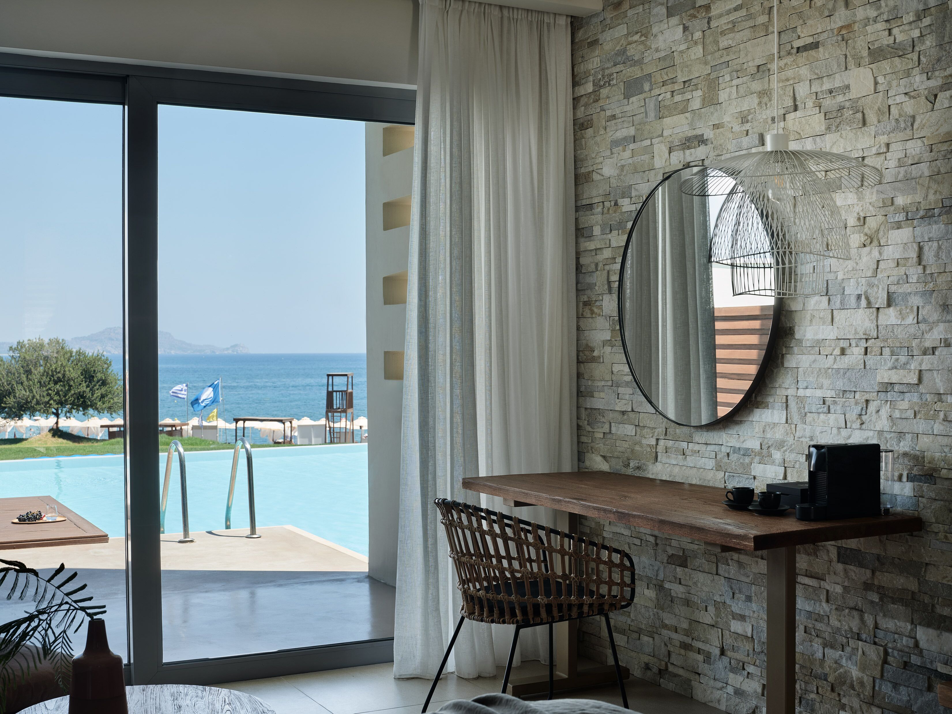 Une pièce avec un mur en pierre, un miroir rond, une table en bois, une chaise en osier et une vue sur une piscine extérieure et une plage.