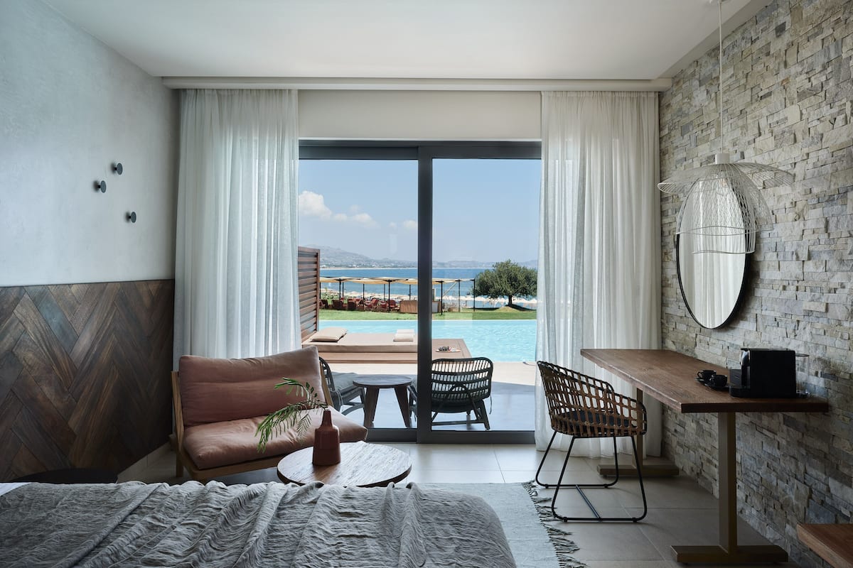 Une chambre d’hôtel moderne dotée d’une grande porte-fenêtre coulissante donnant sur un espace piscine, d’un bureau en bois, d’un canapé confortable et d’une table à manger.