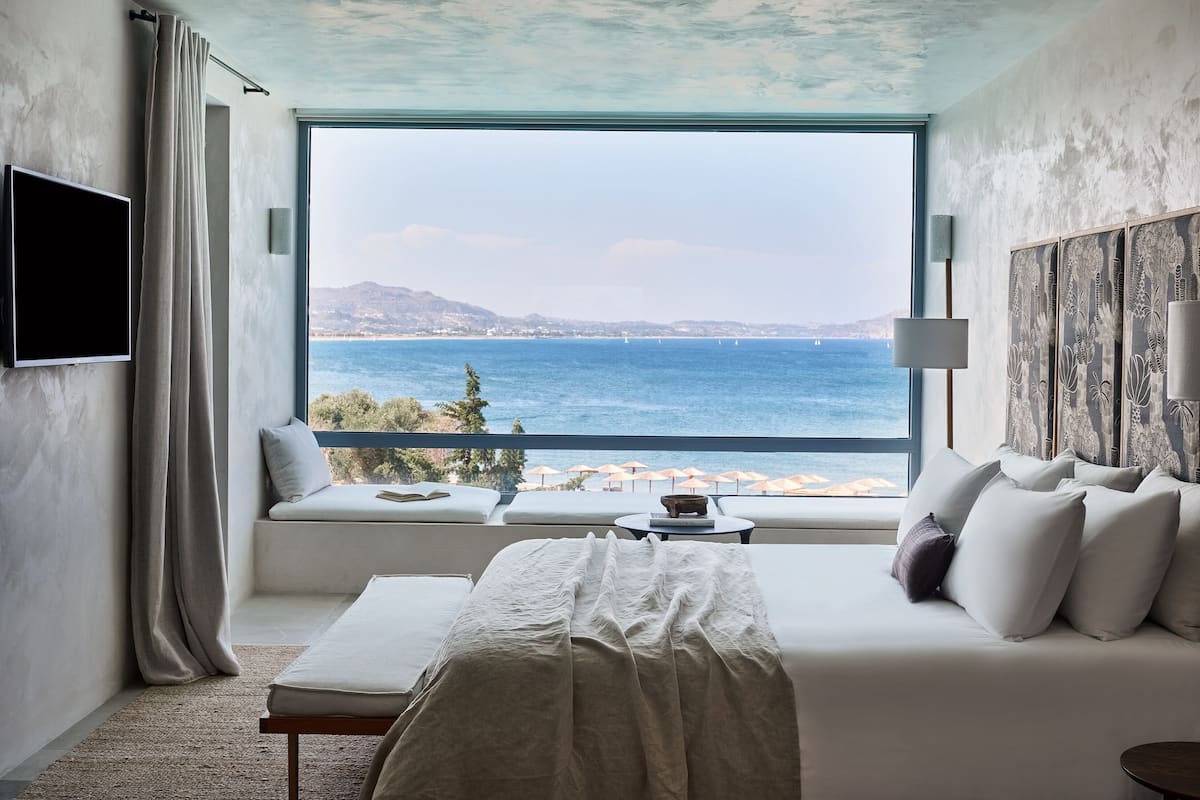 Une chambre d’hôtel dotée d’une grande fenêtre donnant sur la mer, d’un lit, d’une table de chevet et d’un téléviseur à écran plat fixé au mur.