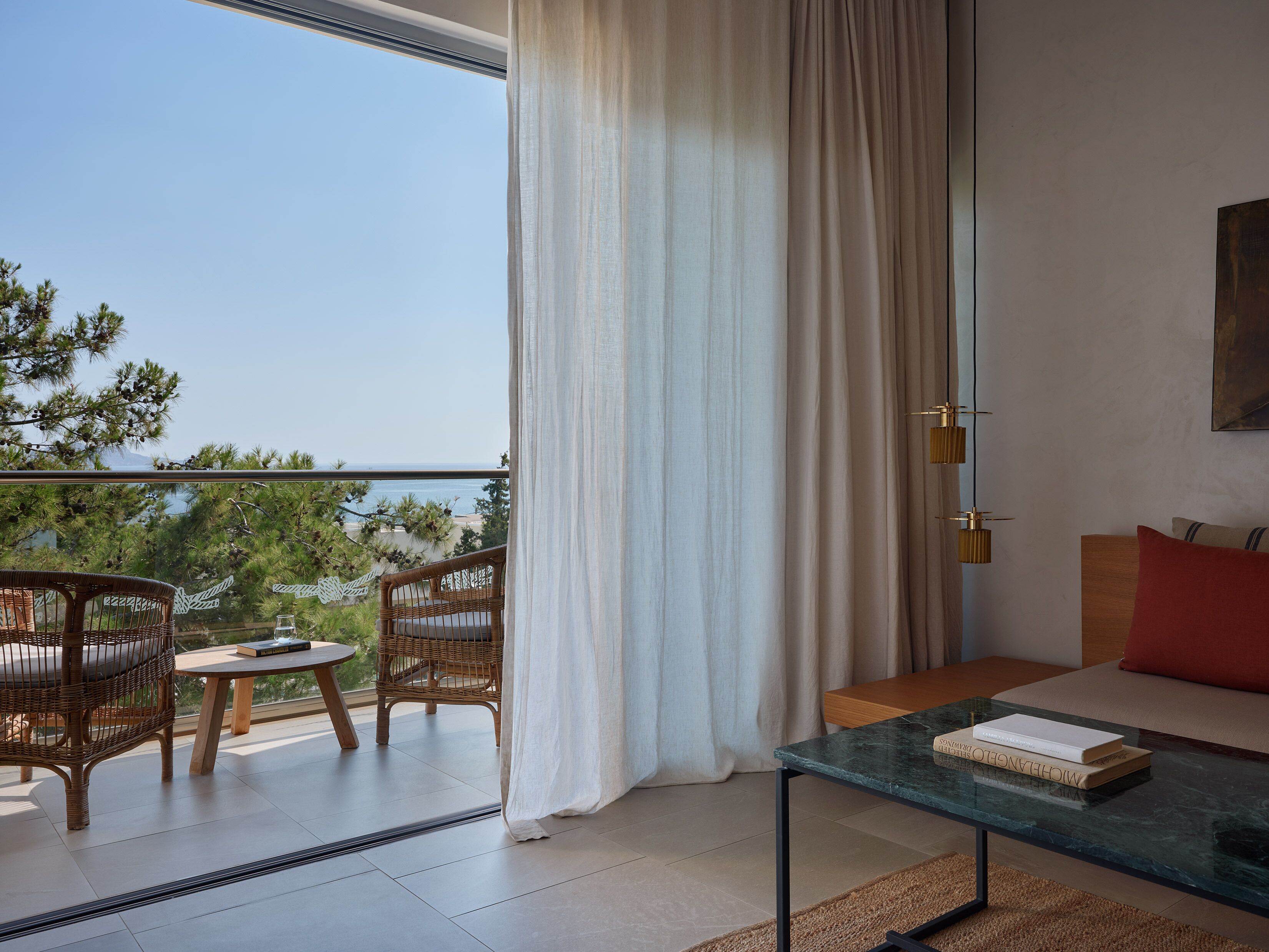 Un balcón con sillas de mimbre y una mesa, una mesita de centro de cristal con libros, y vista al mar.