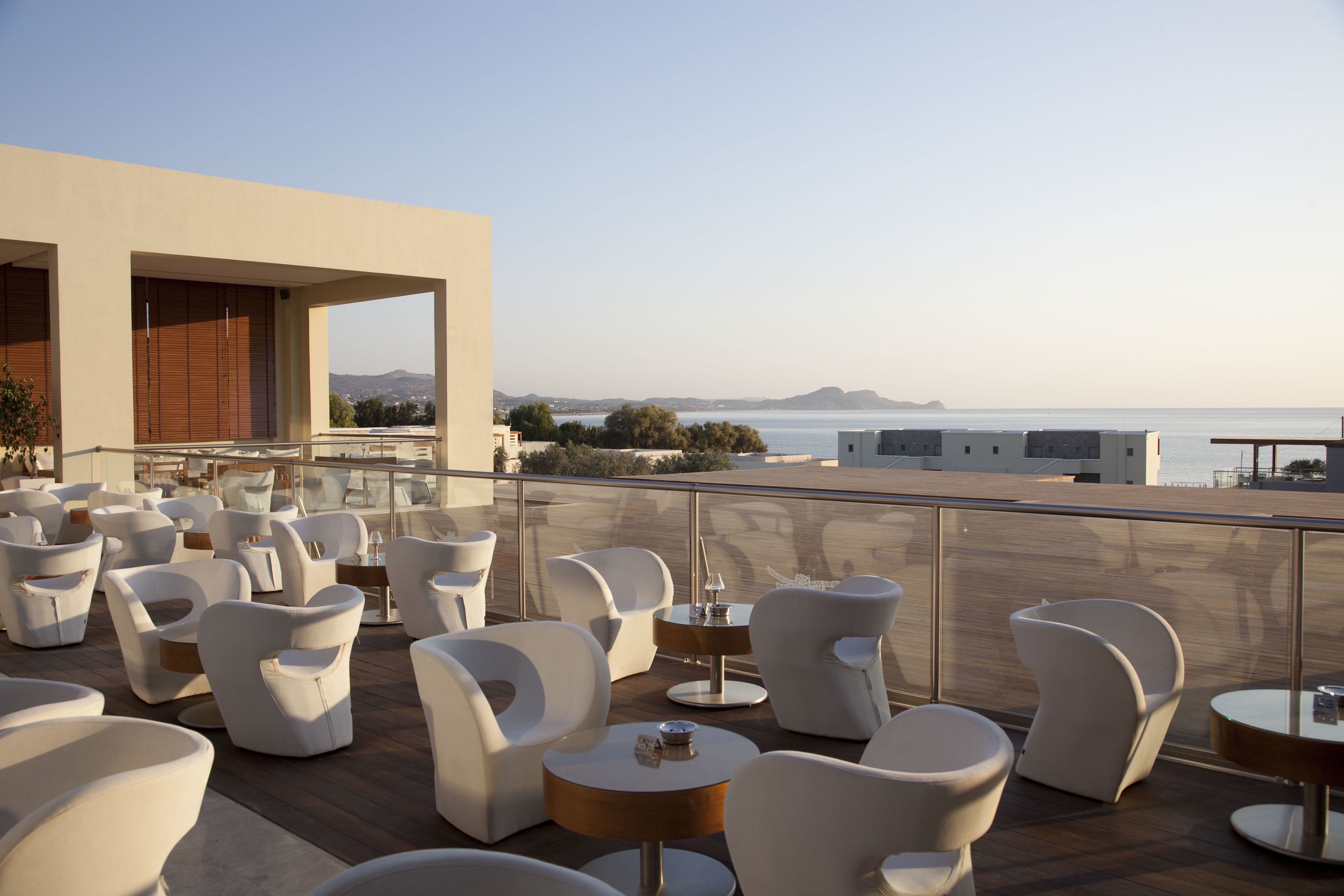 A rooftop terrace with modern white chairs and tables, offering a view of the sea and distant hills.