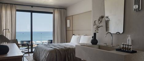 A modern hotel room with a large bed, a desk, a bathroom with a mirror and toiletries, and a view of the ocean.