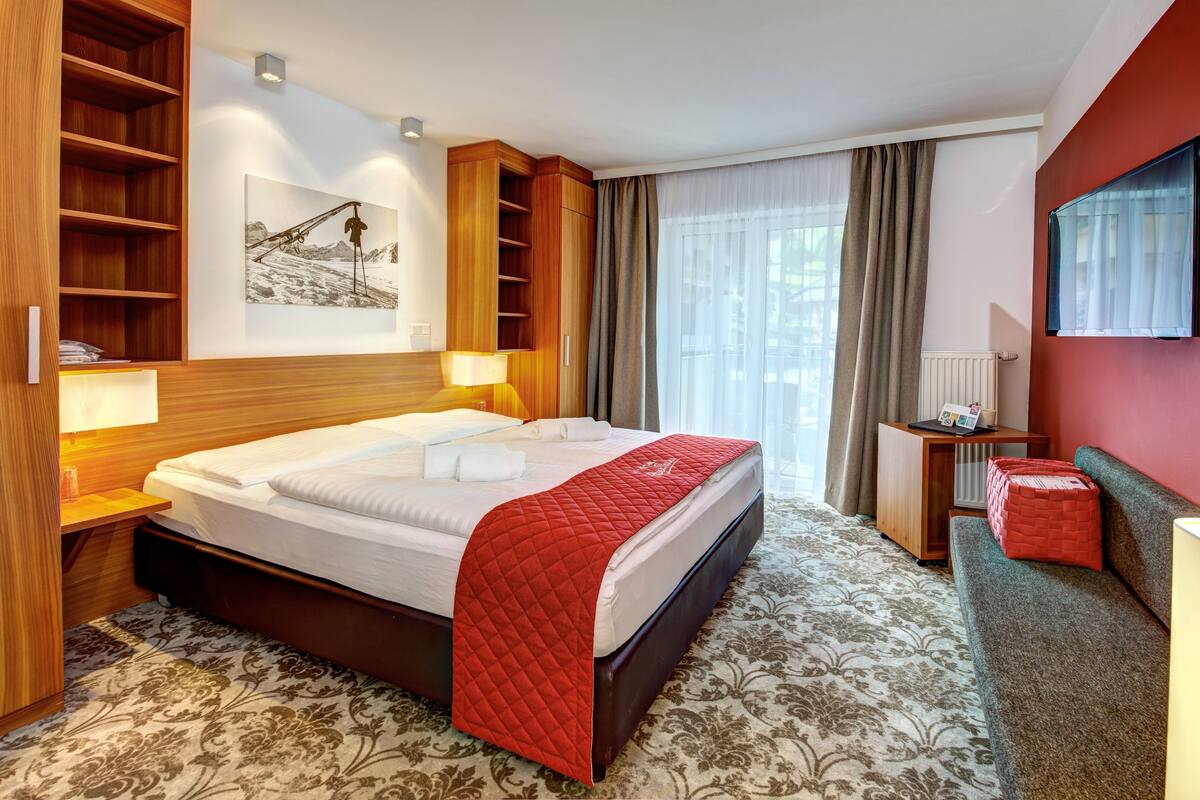 Ein Hotelzimmer mit einem großen Bett, einem Holz-Kopfteil, einer roten gesteppten Tagesdecke, einem kleinen Nachttisch, einem an der Wand befestigten Fernseher und einem Fenster mit Vorhängen.