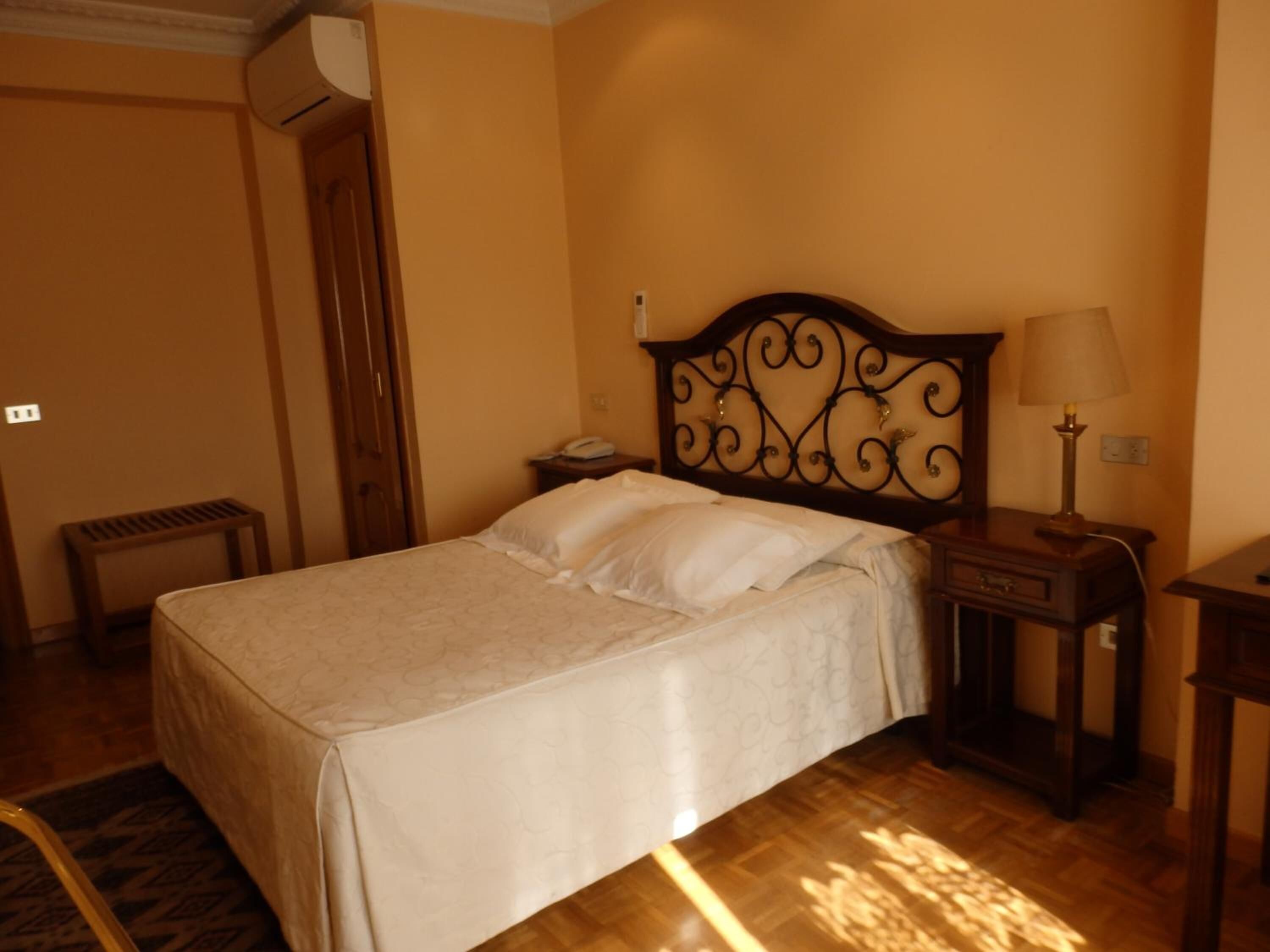 Un lit bien fait, avec une tête de lit, une table de chevet avec une lampe, une petite table de chevet et un appareil de climatisation fixé au mur.