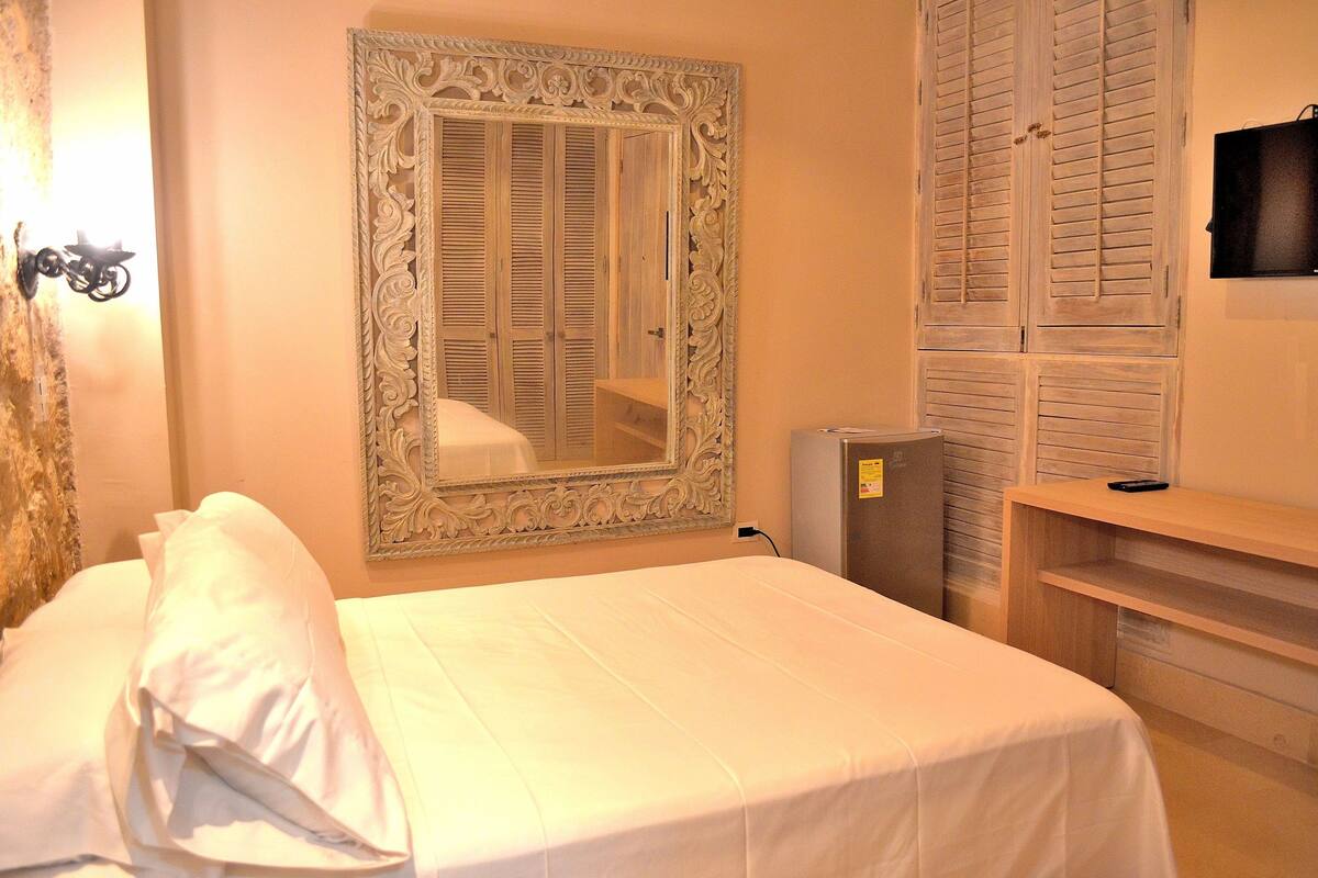 Una habitación de hotel con cama, un espejo de marco decorado, un armario con puertas batientes, un televisor y una mesita.