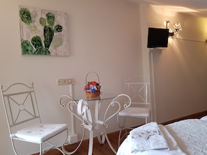 A room with a bed, a white metal chair, a small table with a basket of flowers, a wall-mounted TV, and a cactus painting. - Azabache (Cangas de Onis)