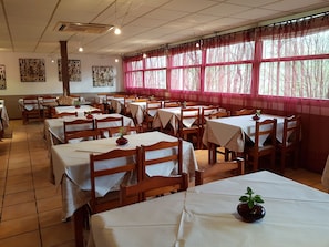 A dining area with multiple tables and chairs, large windows with pink curtains, and wall art. - Azabache (Cangas de Onis)