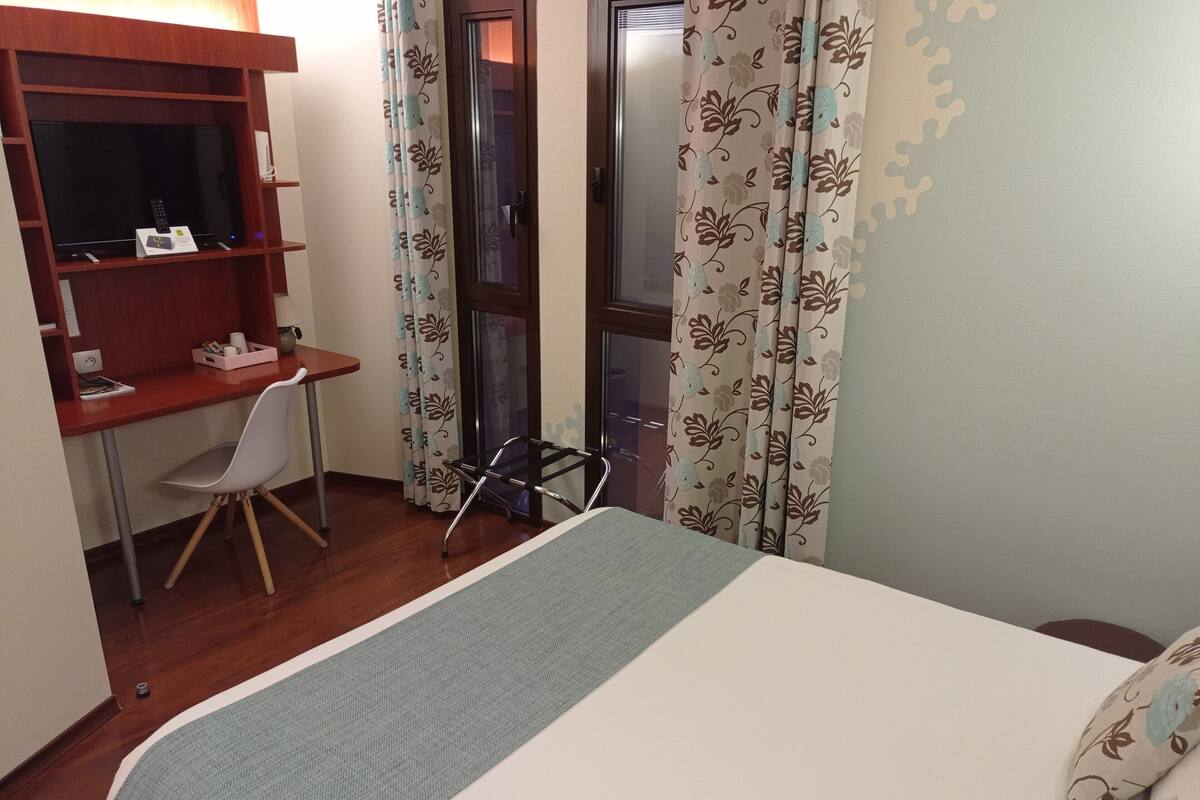 A bedroom with a bed, a desk with a chair, a TV, and a window with curtains.