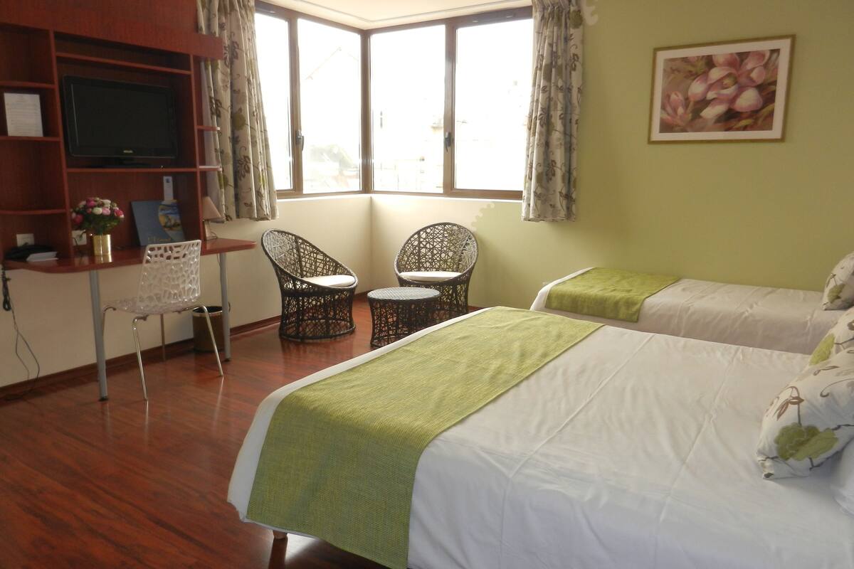 A hotel room with two beds, a desk with a chair, a television, and a large window with curtains.