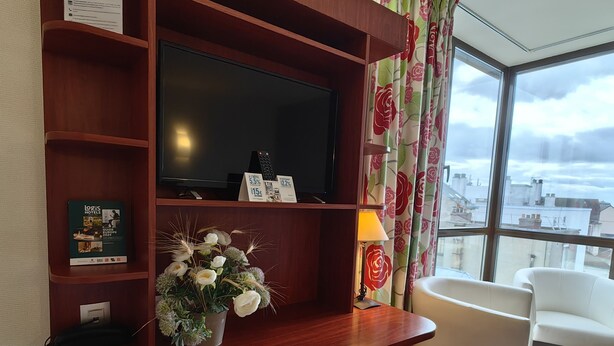 A hotel room with a wooden TV stand, a flat-screen TV, a potted plant, a remote control, and a view of the city through large windows. - L'Hôtel (Chartres)