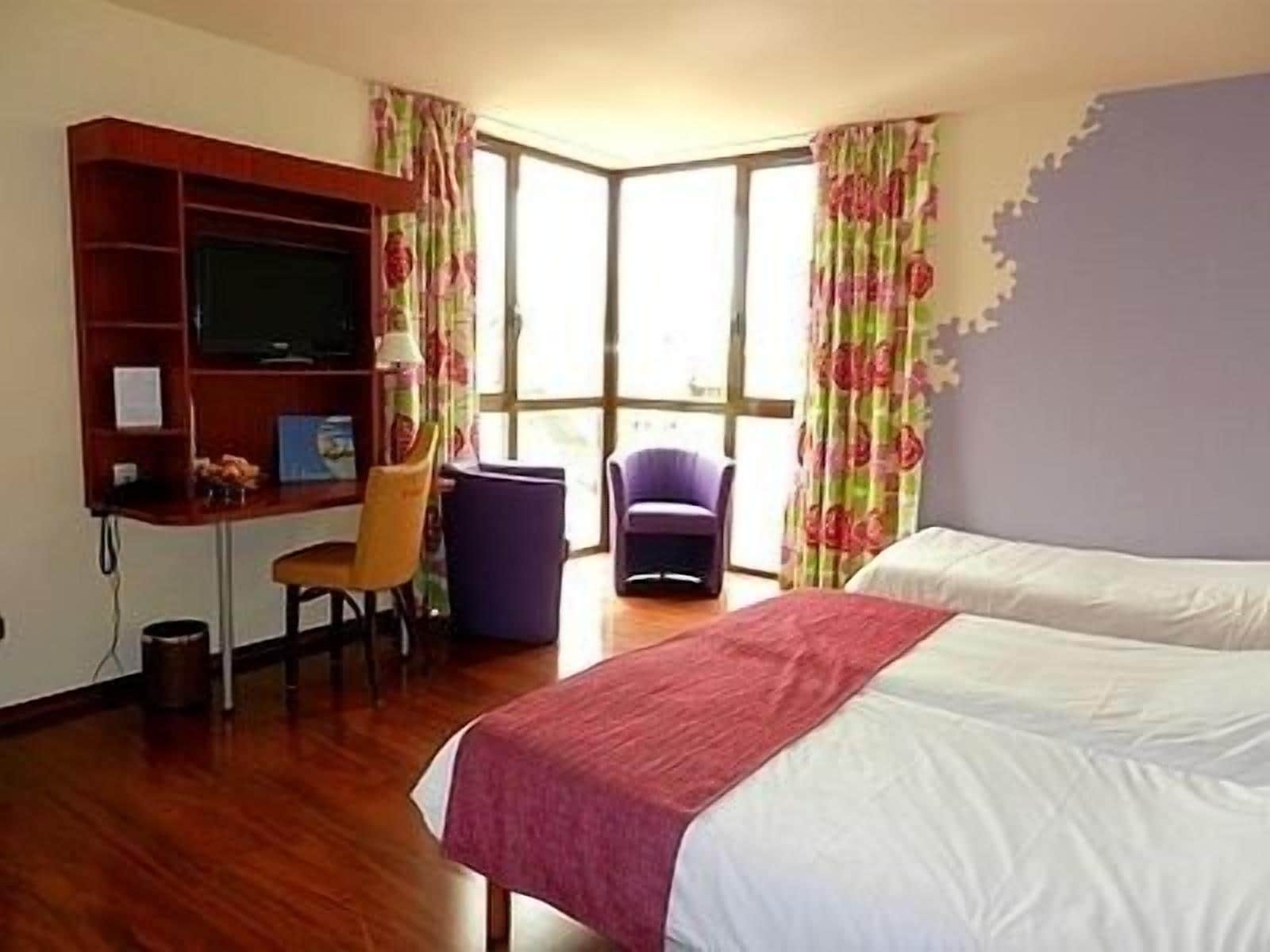 Una habitación de hotel con cama, escritorio, silla, televisión y ventana con cortinas.