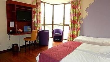 Una habitación de hotel con cama, escritorio, silla, televisión y ventana con cortinas.
