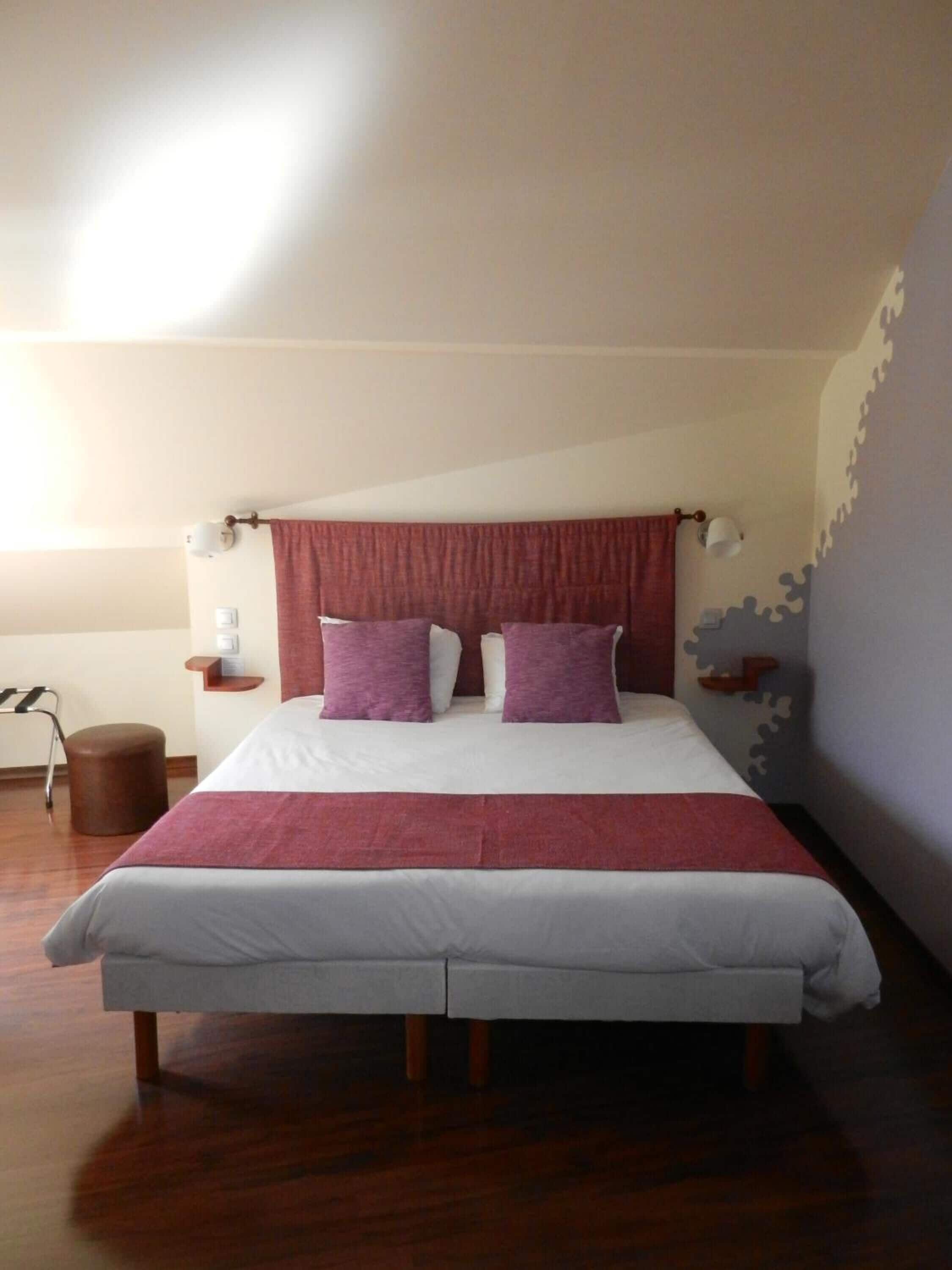 A bedroom with a bed, two pillows, a nightstand, a lamp, a chair, and a wall with a unique design.
