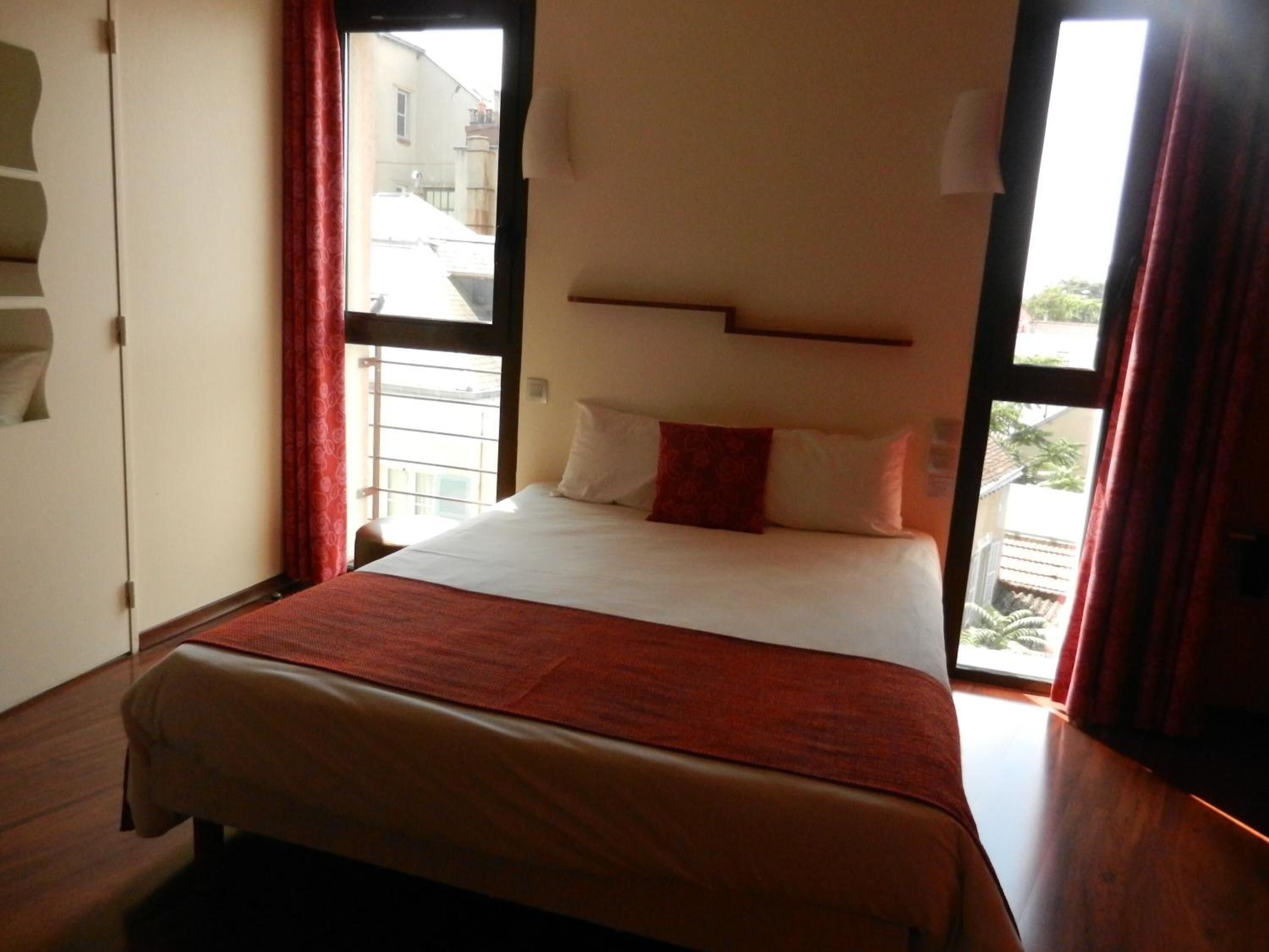 A hotel room with a large bed, a window with a view of buildings, and a red curtain.