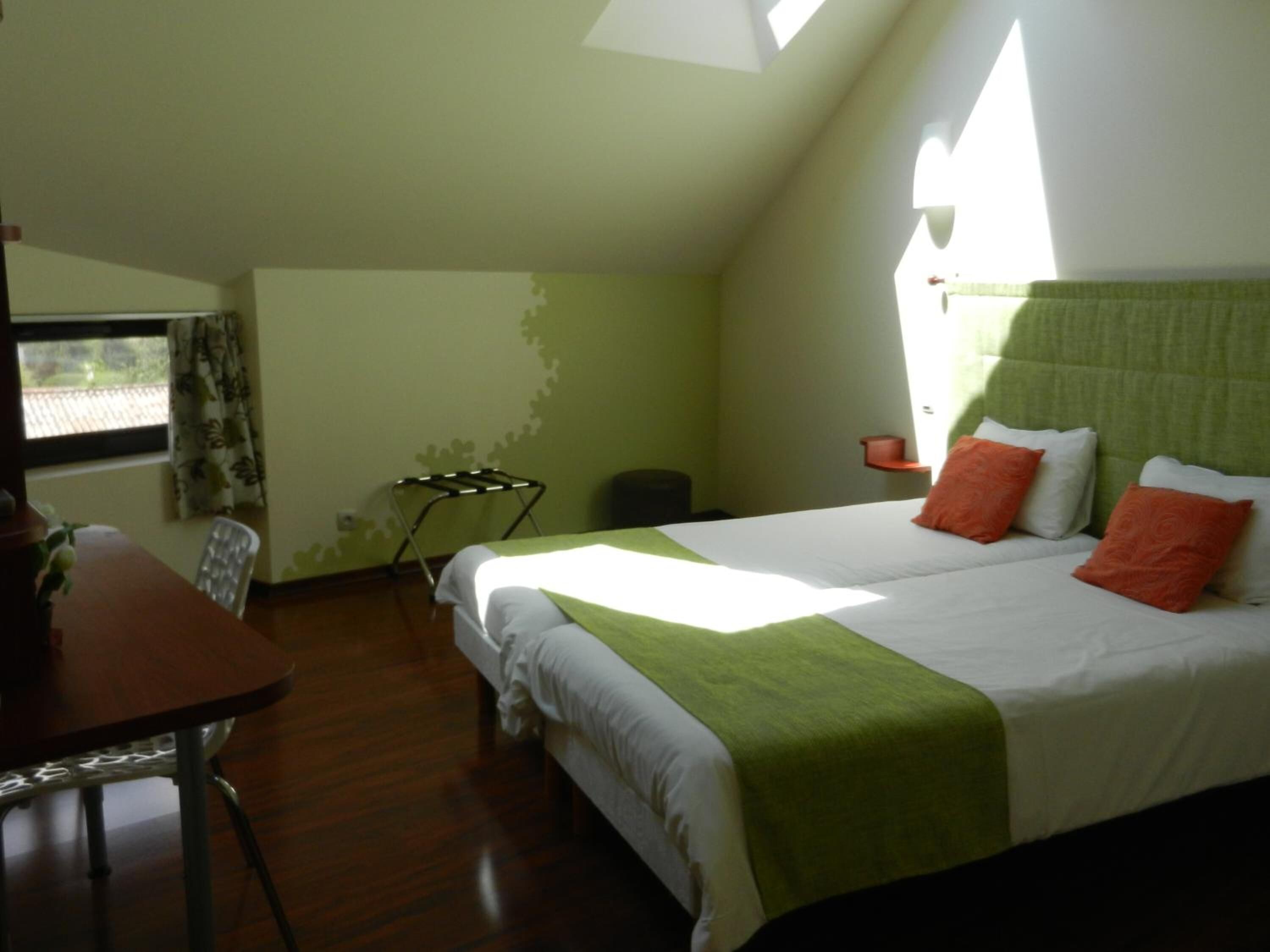 A bedroom with a bed, a table, and a chair. There is a window with curtains, a drying rack, and a small table with a lamp.