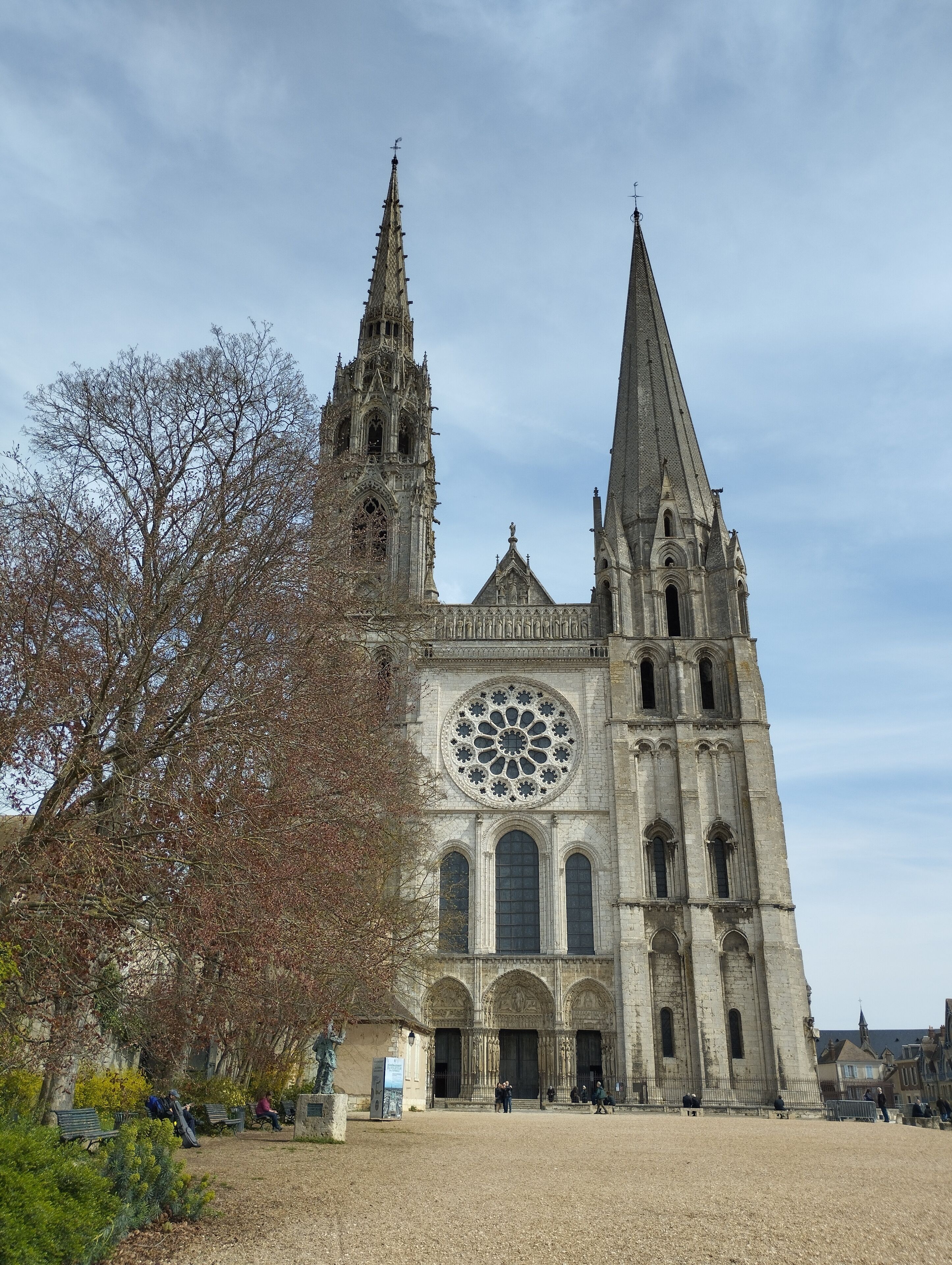 Uma catedral histórica com duas torres imponentes, um vitral rosáceo grande e um pátio com bancos.