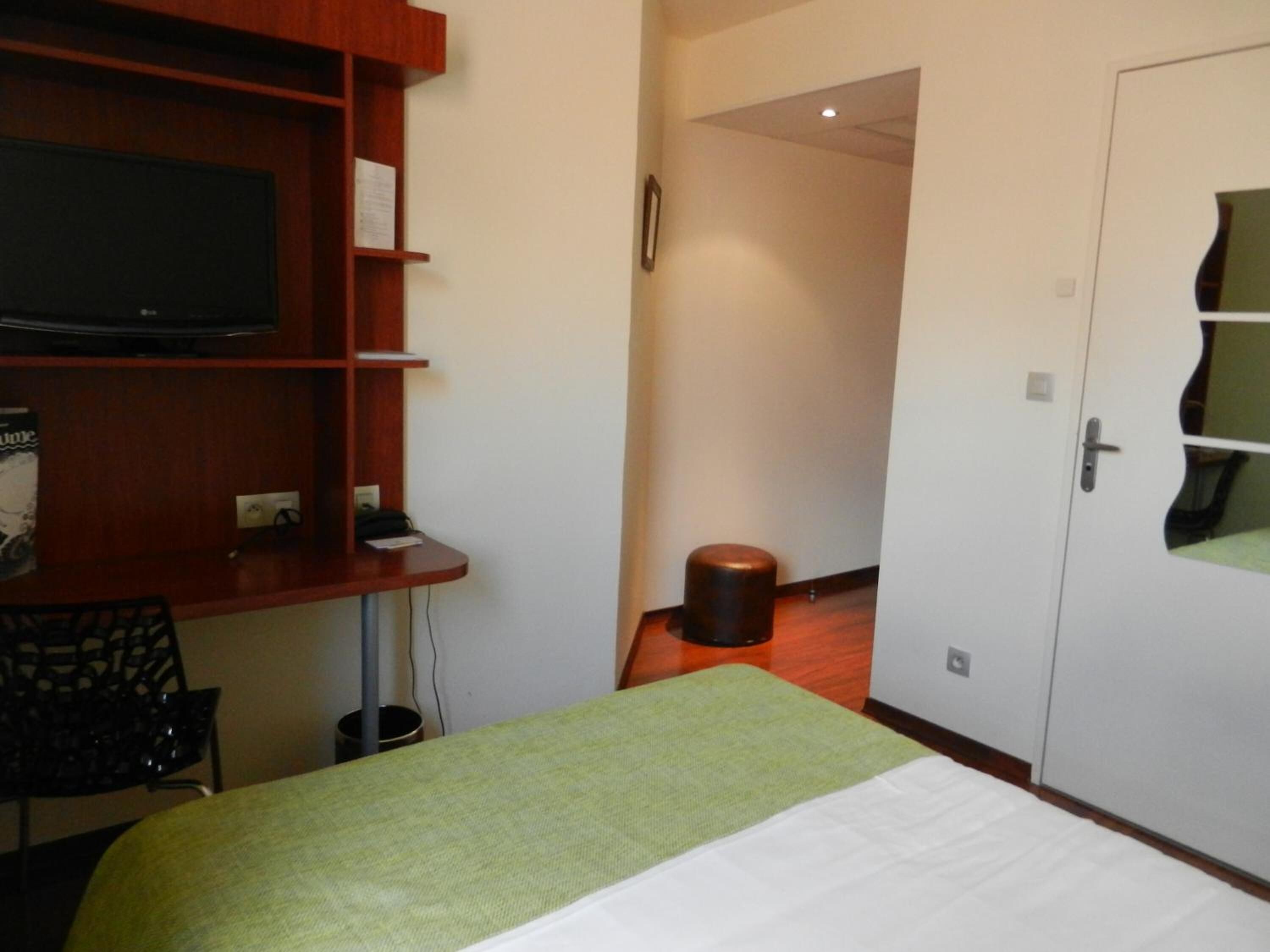 A hotel room with a bed, a TV on a wooden shelf, a desk, and a door with a mirror.