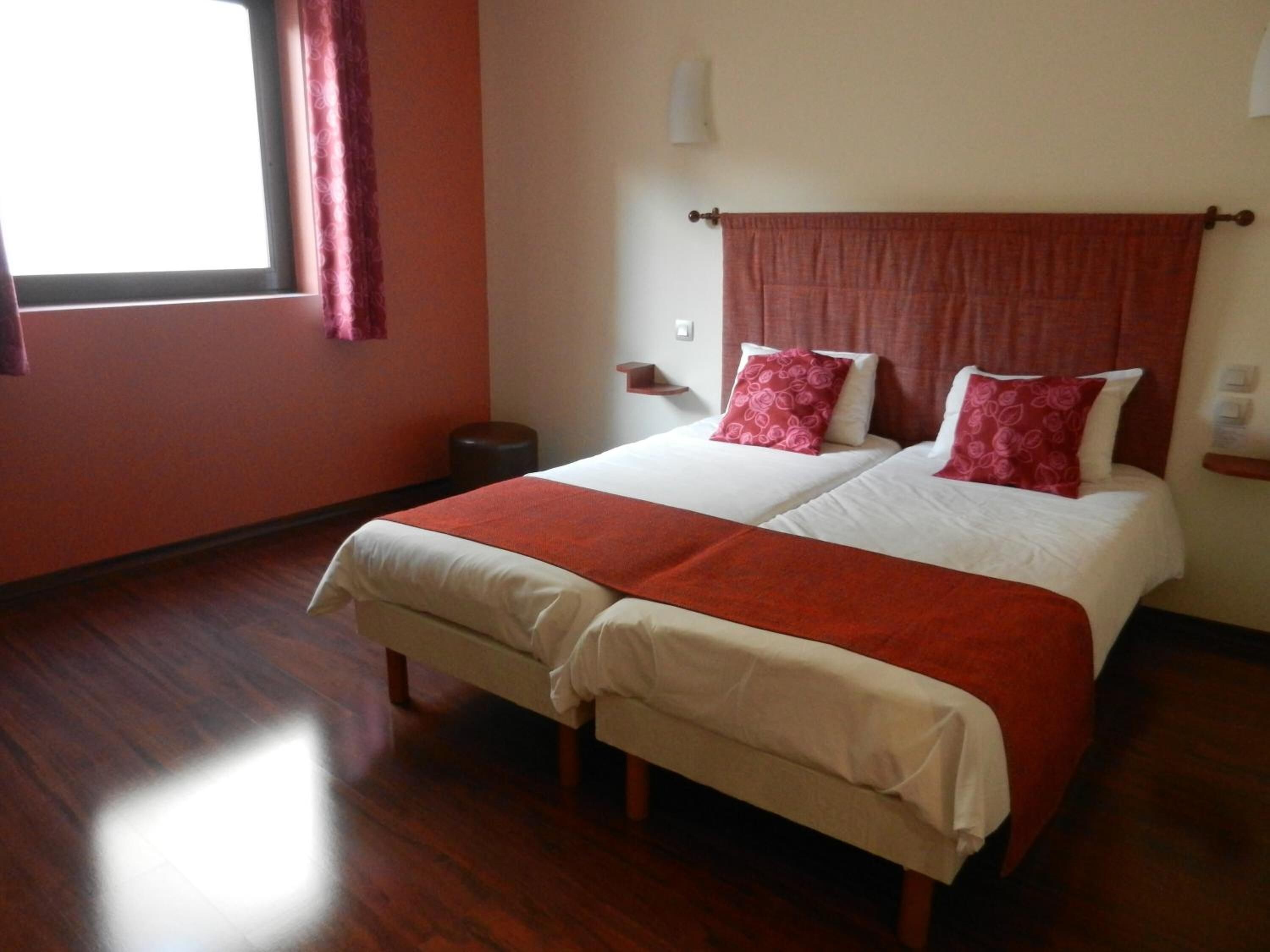 A double bed with a red and white bedspread, a wooden floor, a window with a red curtain, and a wall-mounted light fixture.
