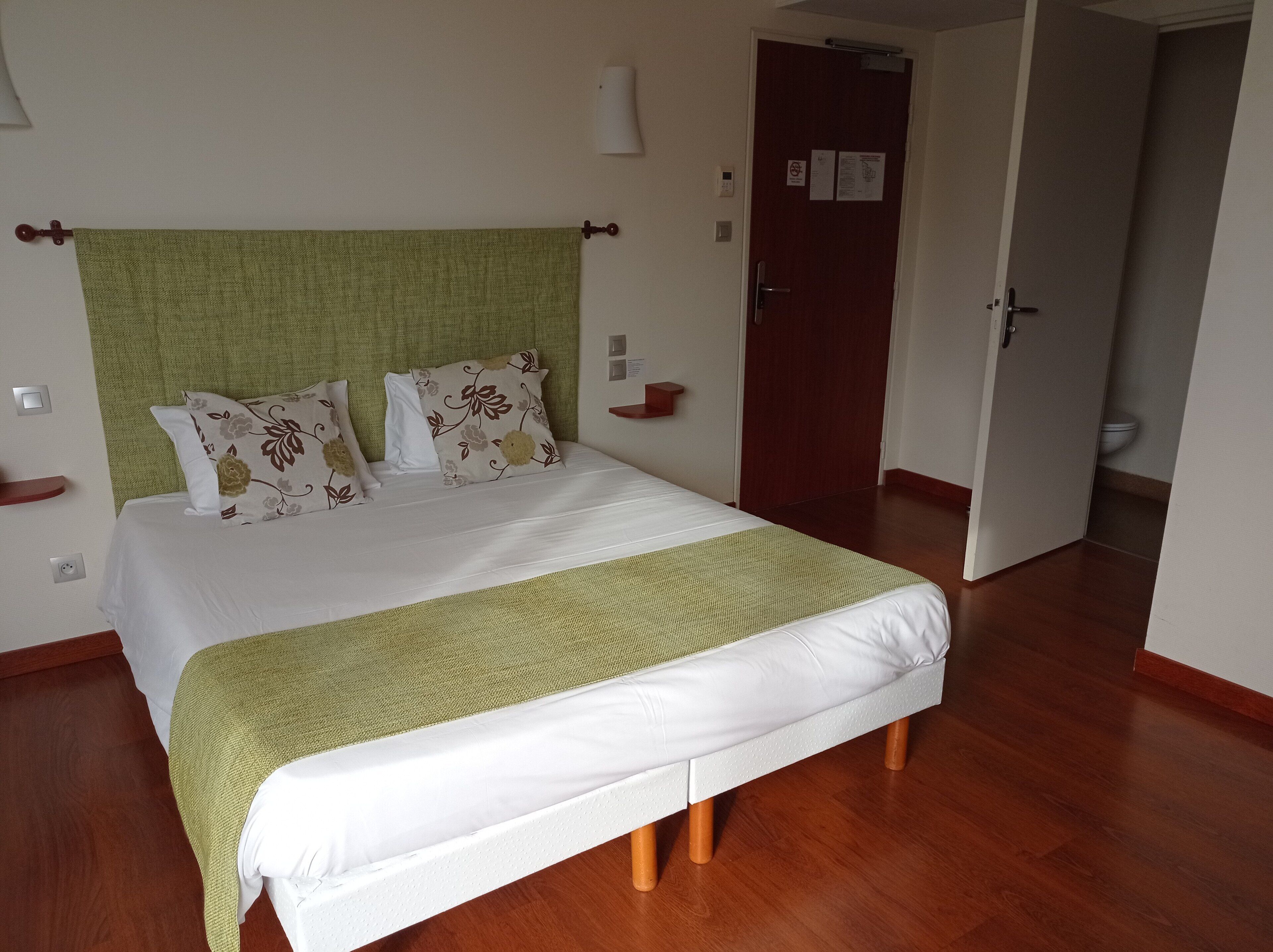 Una cama bien tendida con sábanas blancas y detalles verdes, suelo de madera y una puerta al fondo.