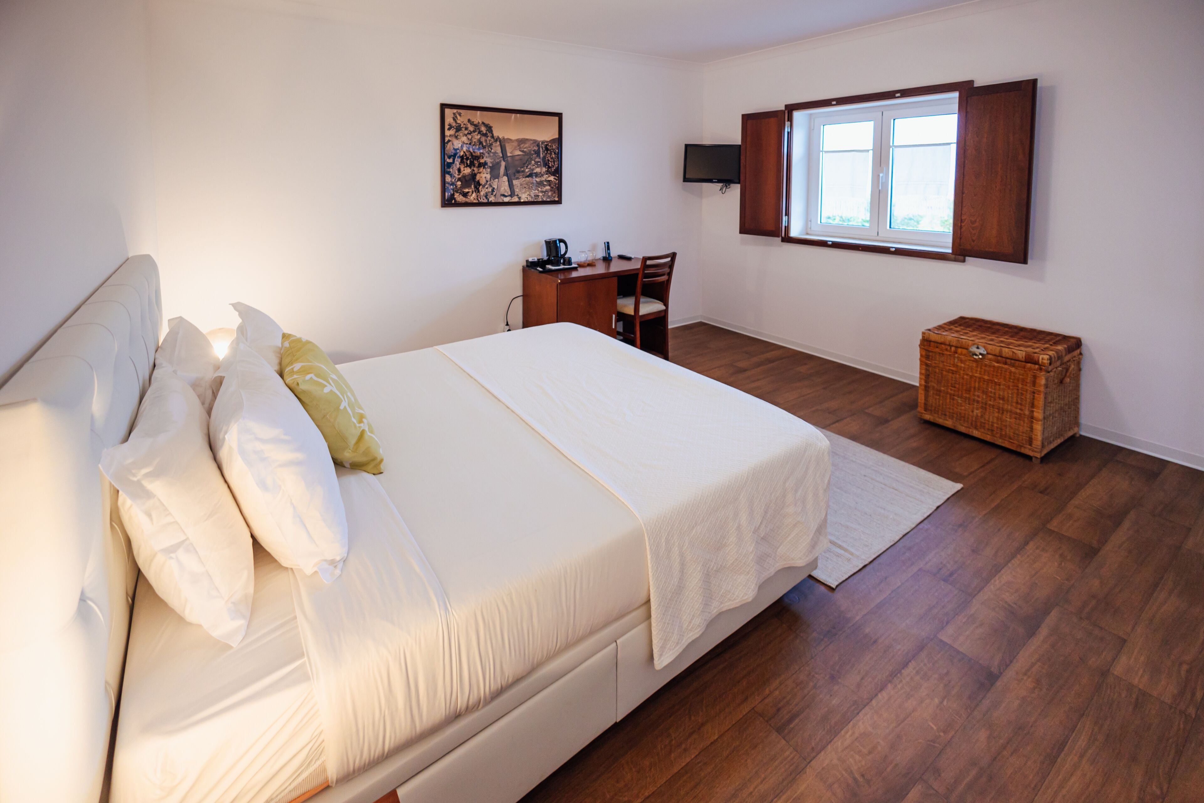 A bedroom with a bed, a desk, a chair, a television, a window, and a small wooden chest.