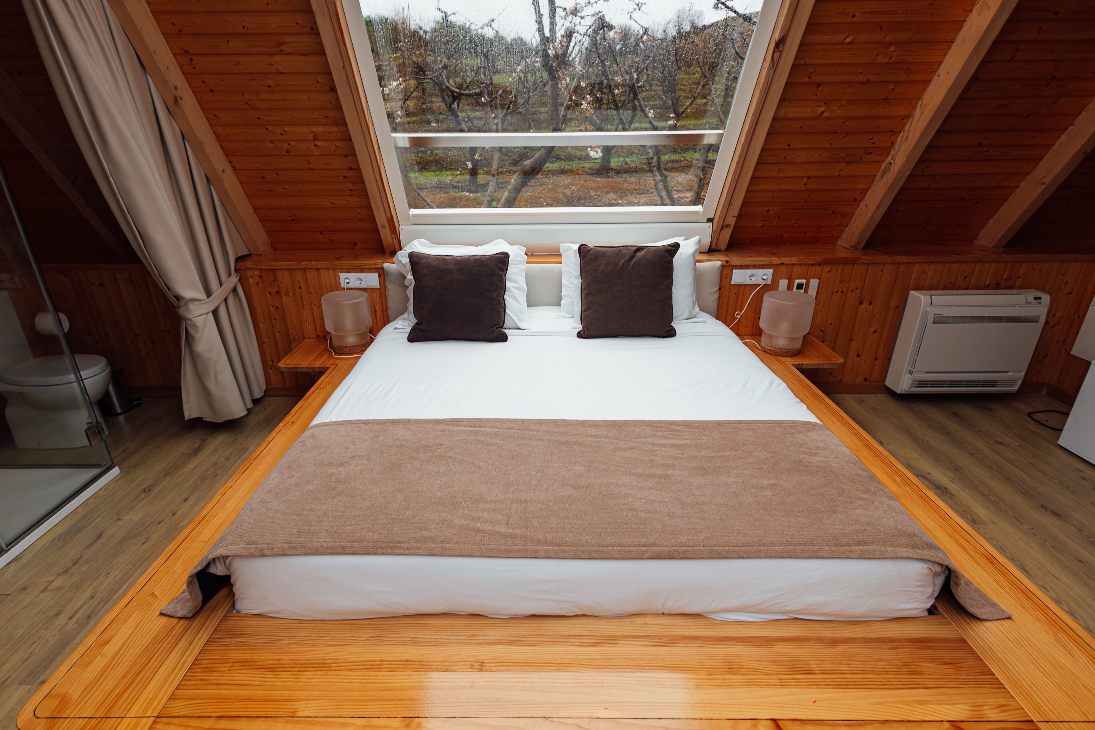 A bedroom with a large bed, wooden walls, a window with a view of trees, and a built-in heater.