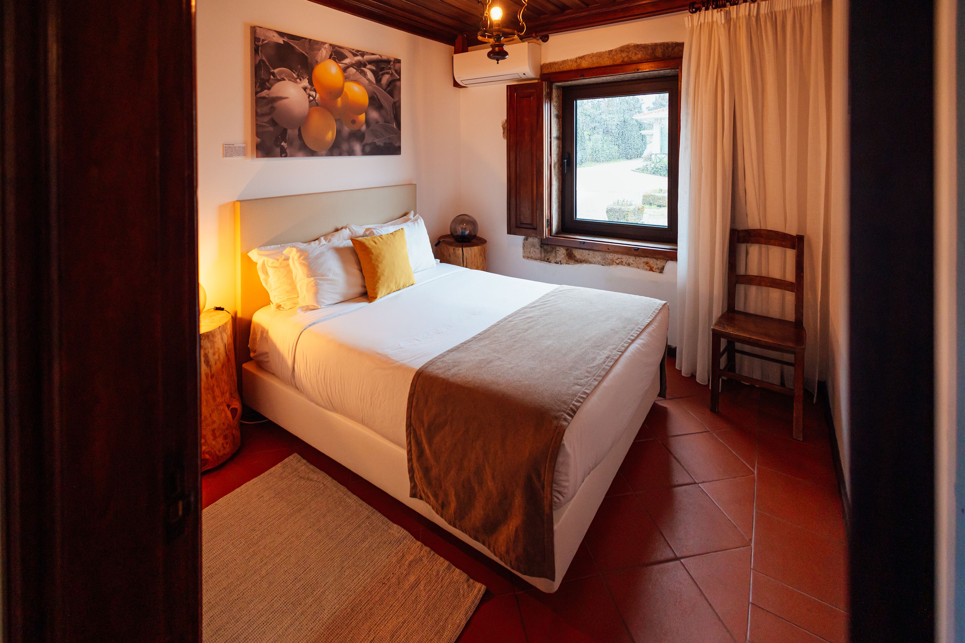 Une chambre avec un grand lit, une table de chevet, une chaise, une fenêtre avec des rideaux et un tableau au mur.