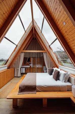 A triangular wooden room with a bed, a bathroom, and large windows.