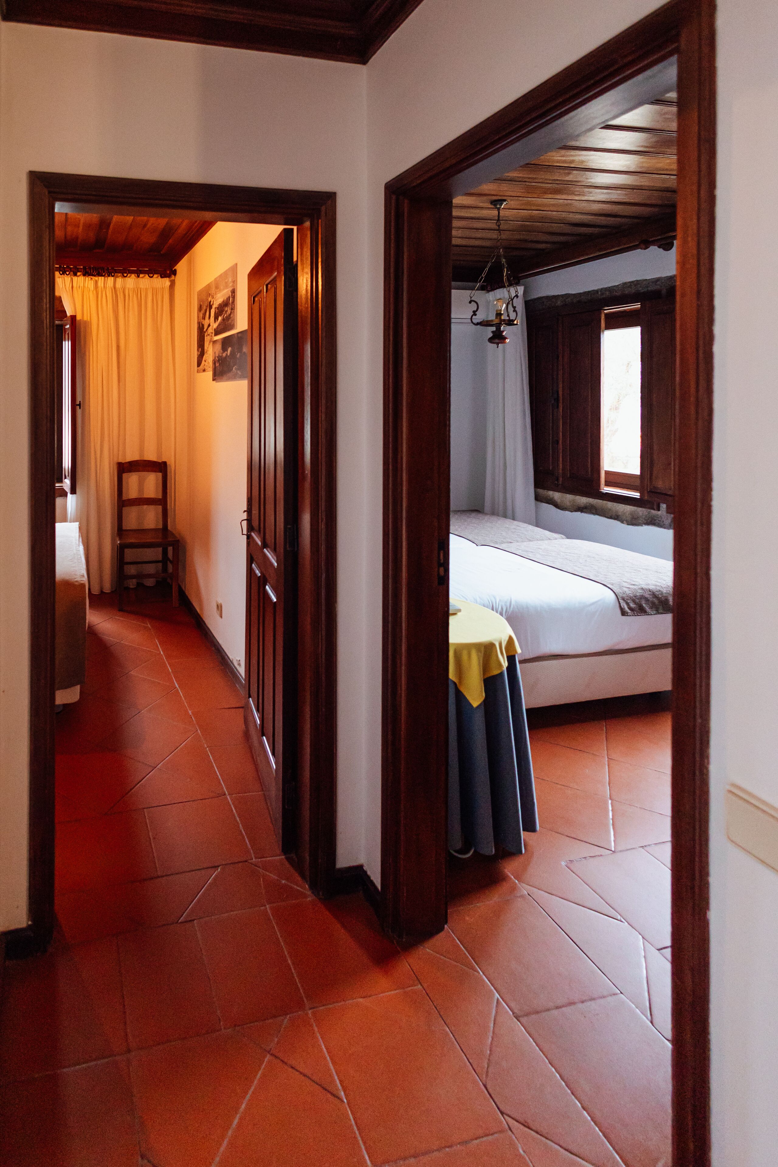 Un couloir étroit, avec un lit au fond, une chaise et une table recouverte d’une nappe jaune.