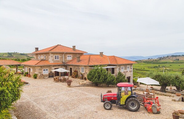 A stone house with a tiled roof, a paved courtyard, and a red tractor parked in front.
