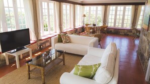A spacious living room with a large flat-screen TV, a glass coffee table, and a comfortable sofa. There are wicker chairs and a small table with a lamp. The room has large windows with curtains, allowing natural light to fill the space. - Quinta da Barroca (Armamar)