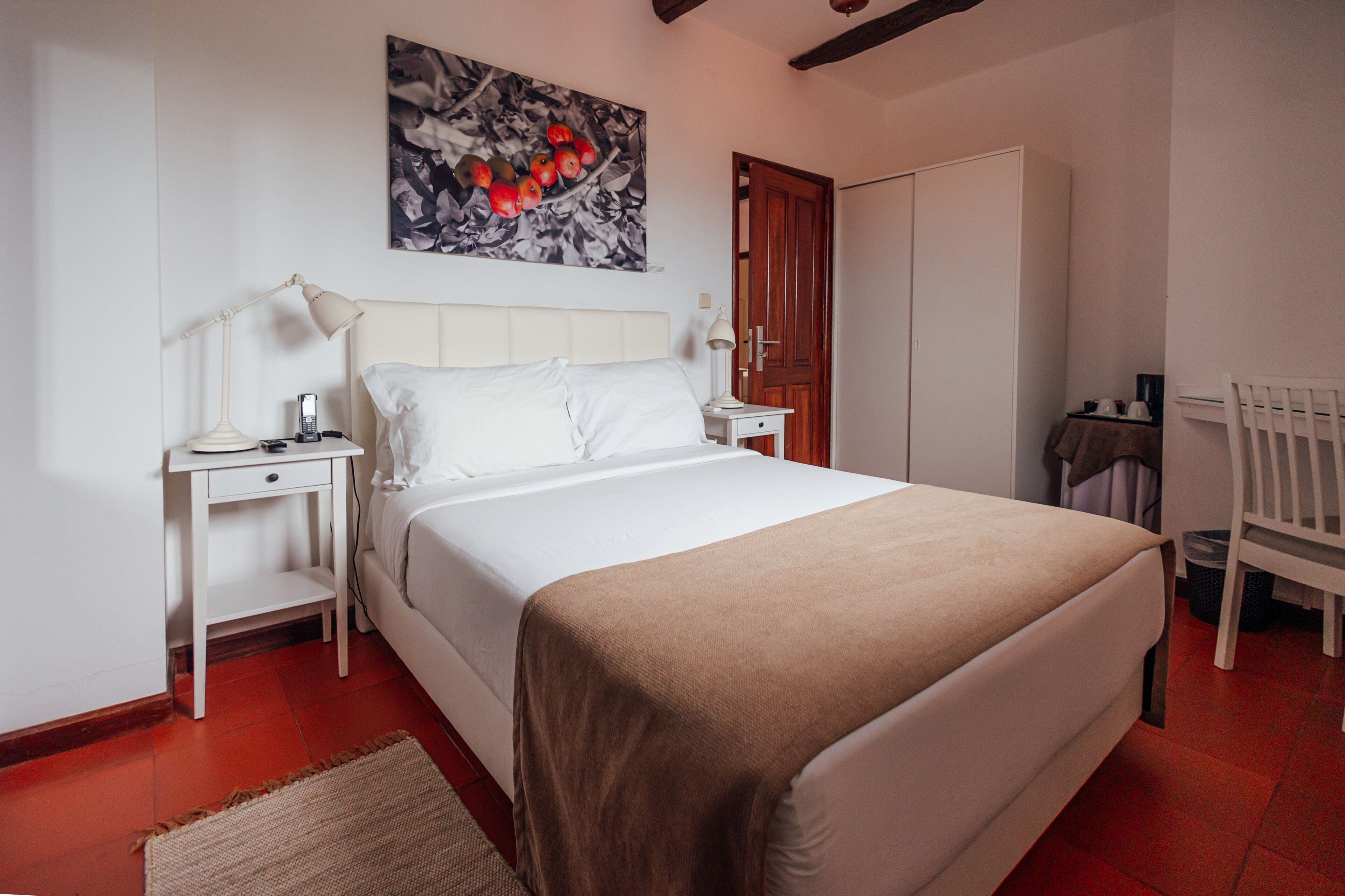 Un dormitorio con cama, mesitas de noche, lámpara, silla, puerta y un cuadro en la pared.