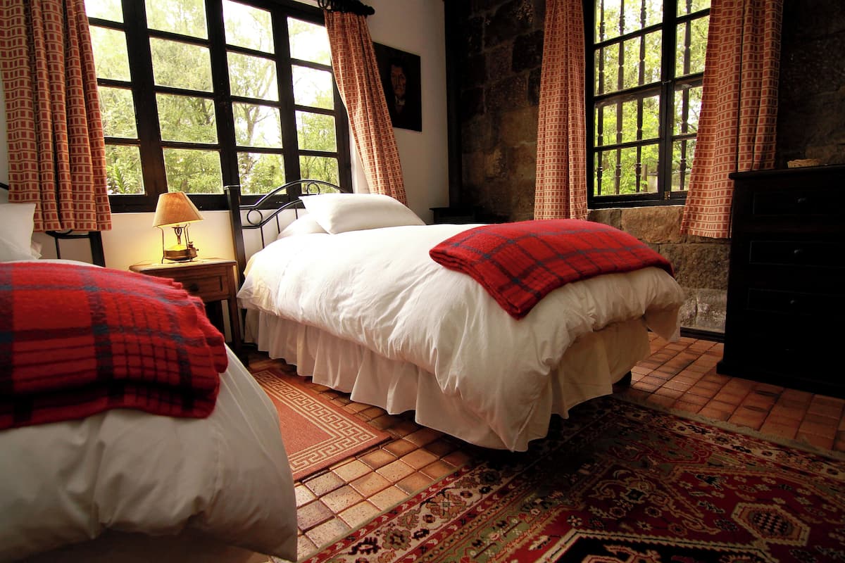 A bedroom with two beds, a nightstand with a lamp, a dresser, and large windows with curtains.