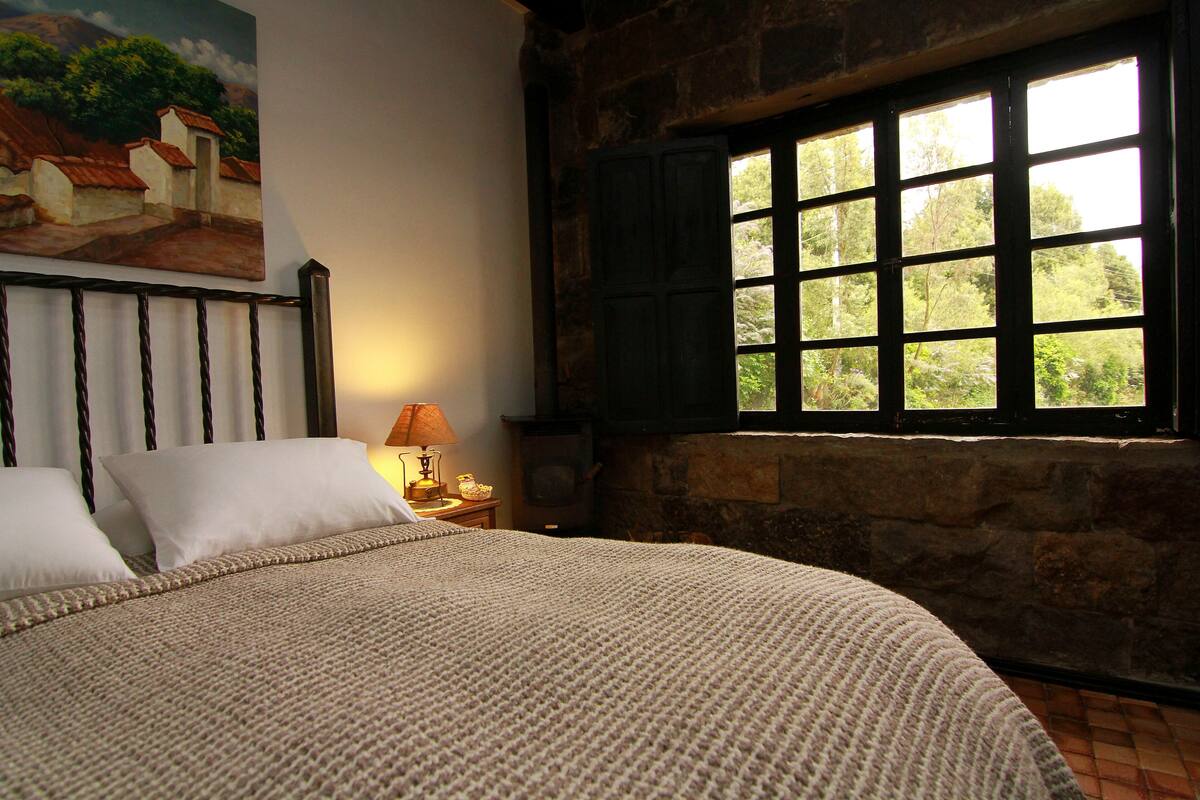 A bed with a knitted blanket, a painting on the wall, a window with shutters, and a nightstand with a lamp.