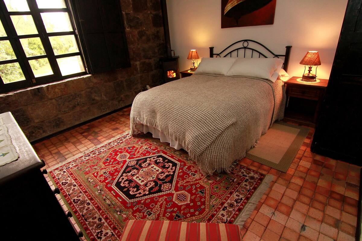A bedroom with a bed, two bedside lamps, a rug, and a window with a view of greenery.